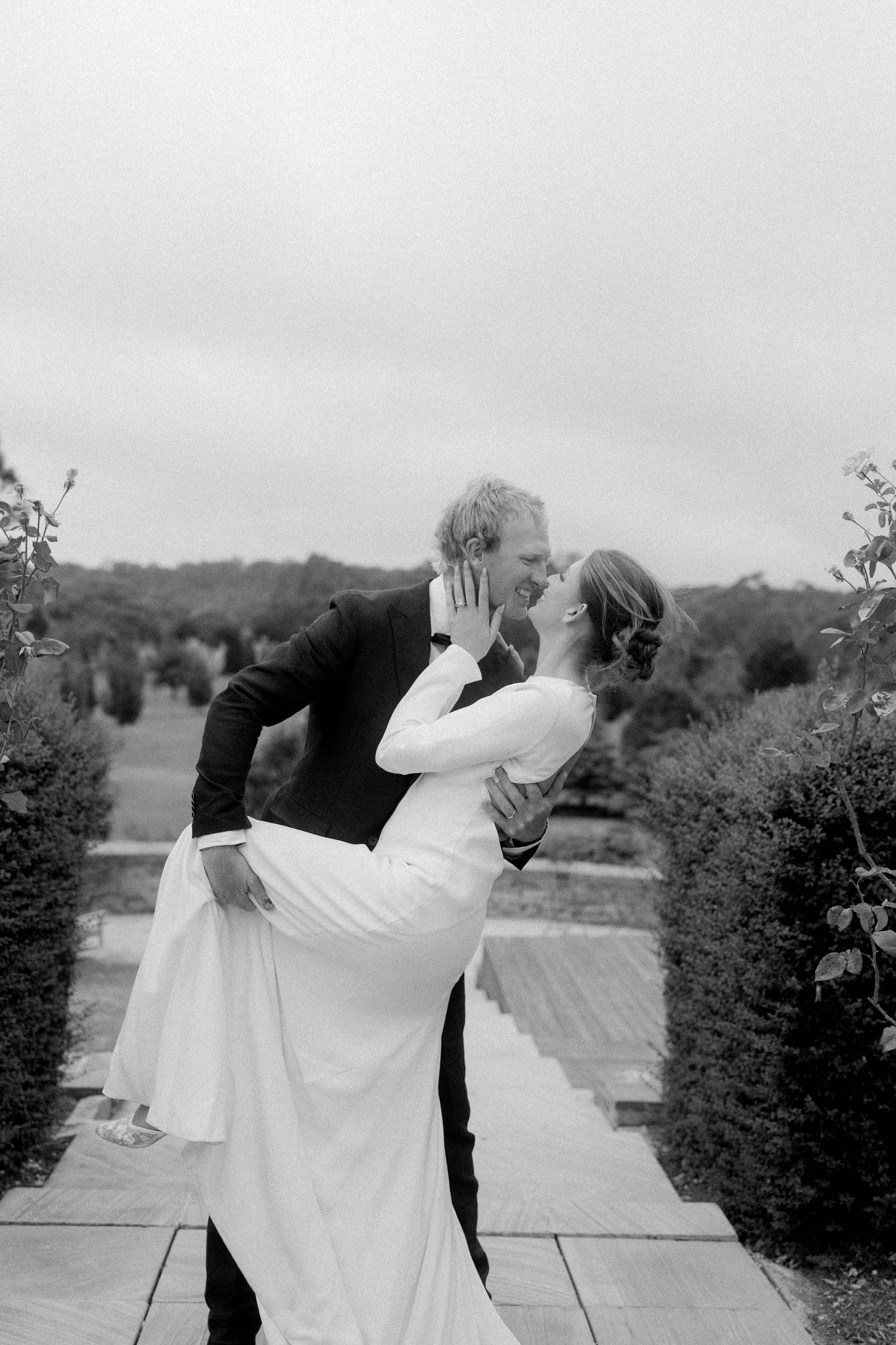 Black and white photo of groom lifting bride for a romantic kiss in a manicured garden.