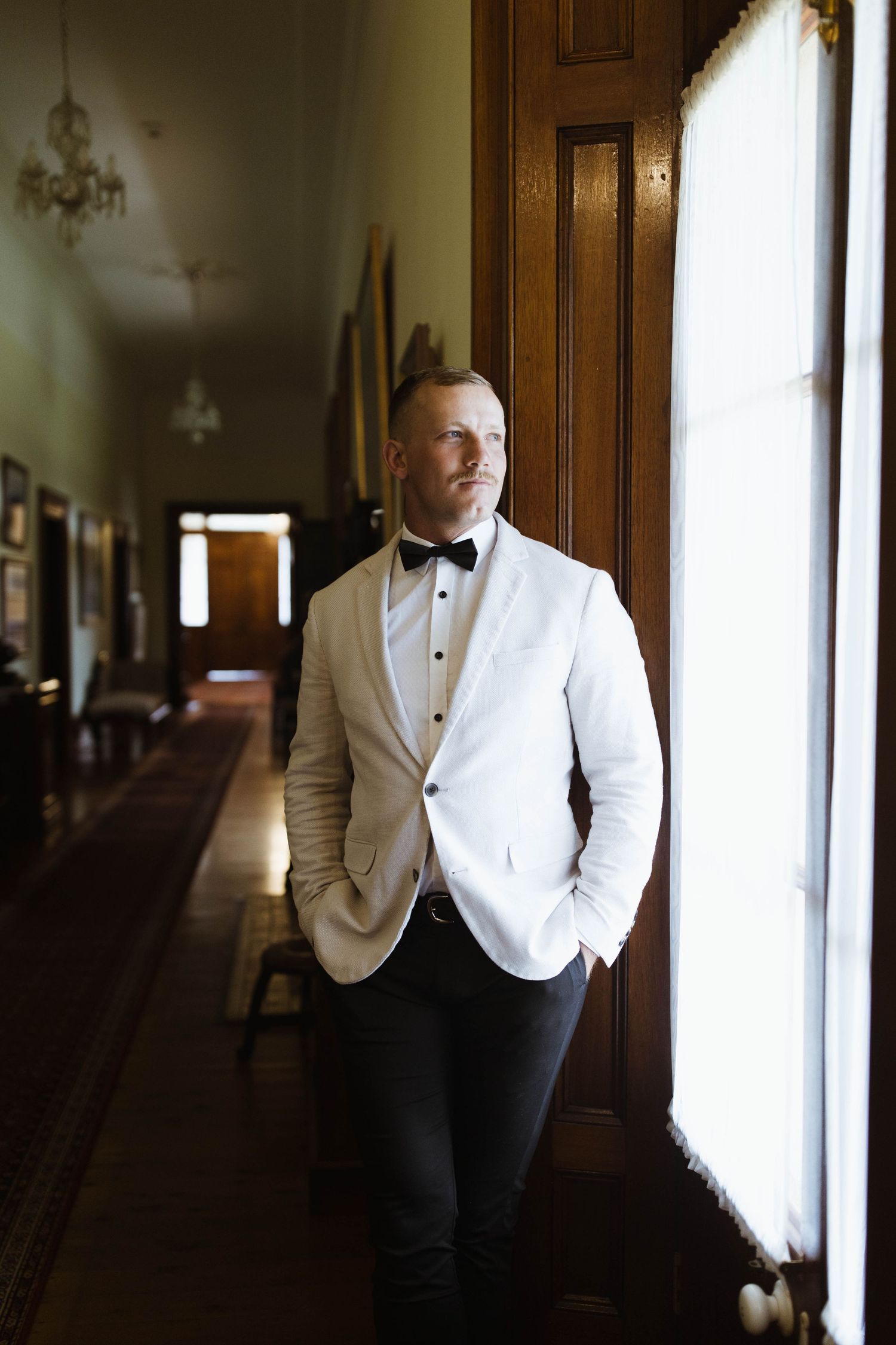 A groom in a white tuxedo jacket and black pants stands near a window in an elegant hallway.