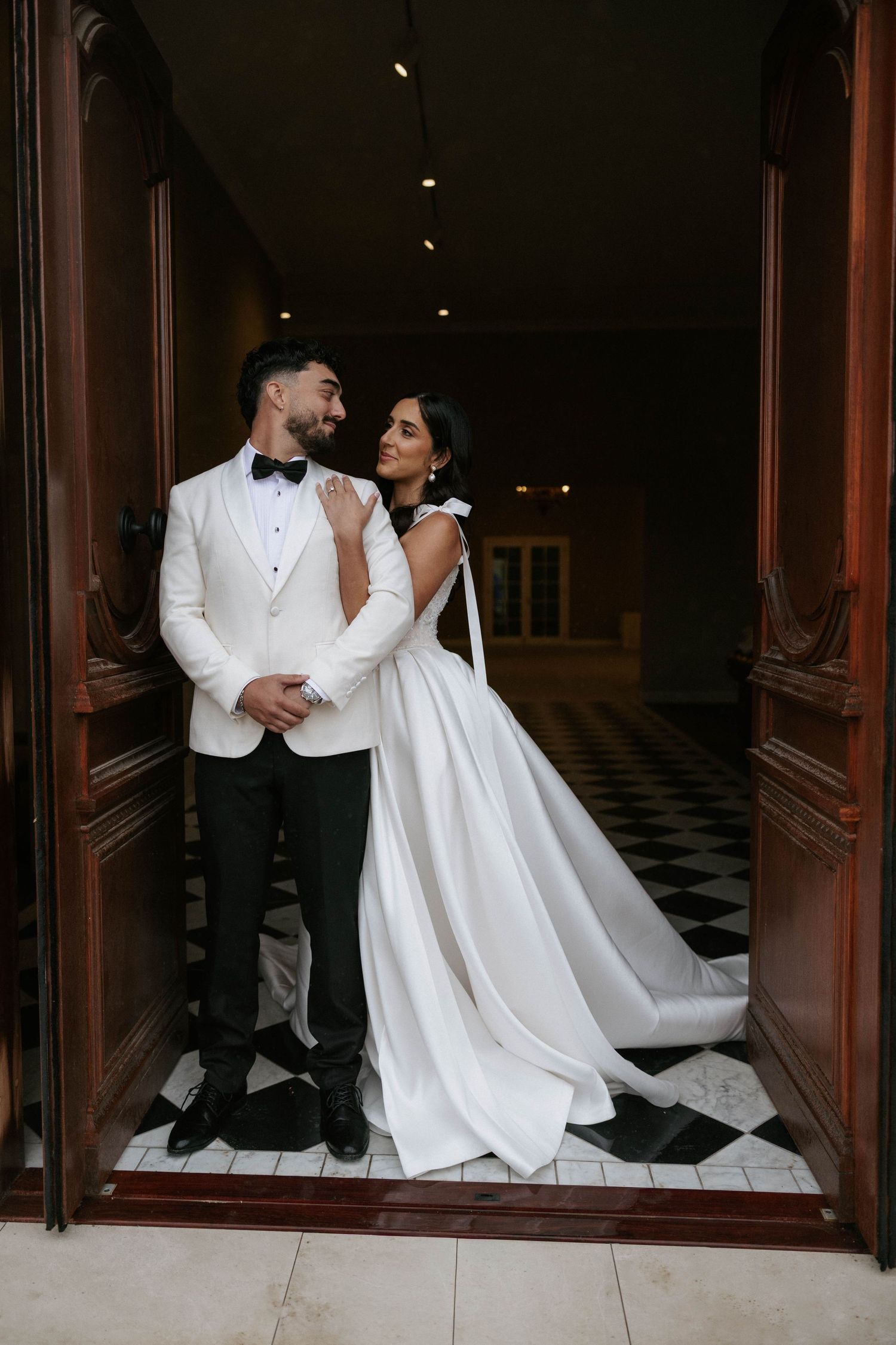 Elegant bride and groom in white attire embrace in ornate doorway with black and white checkered floor.