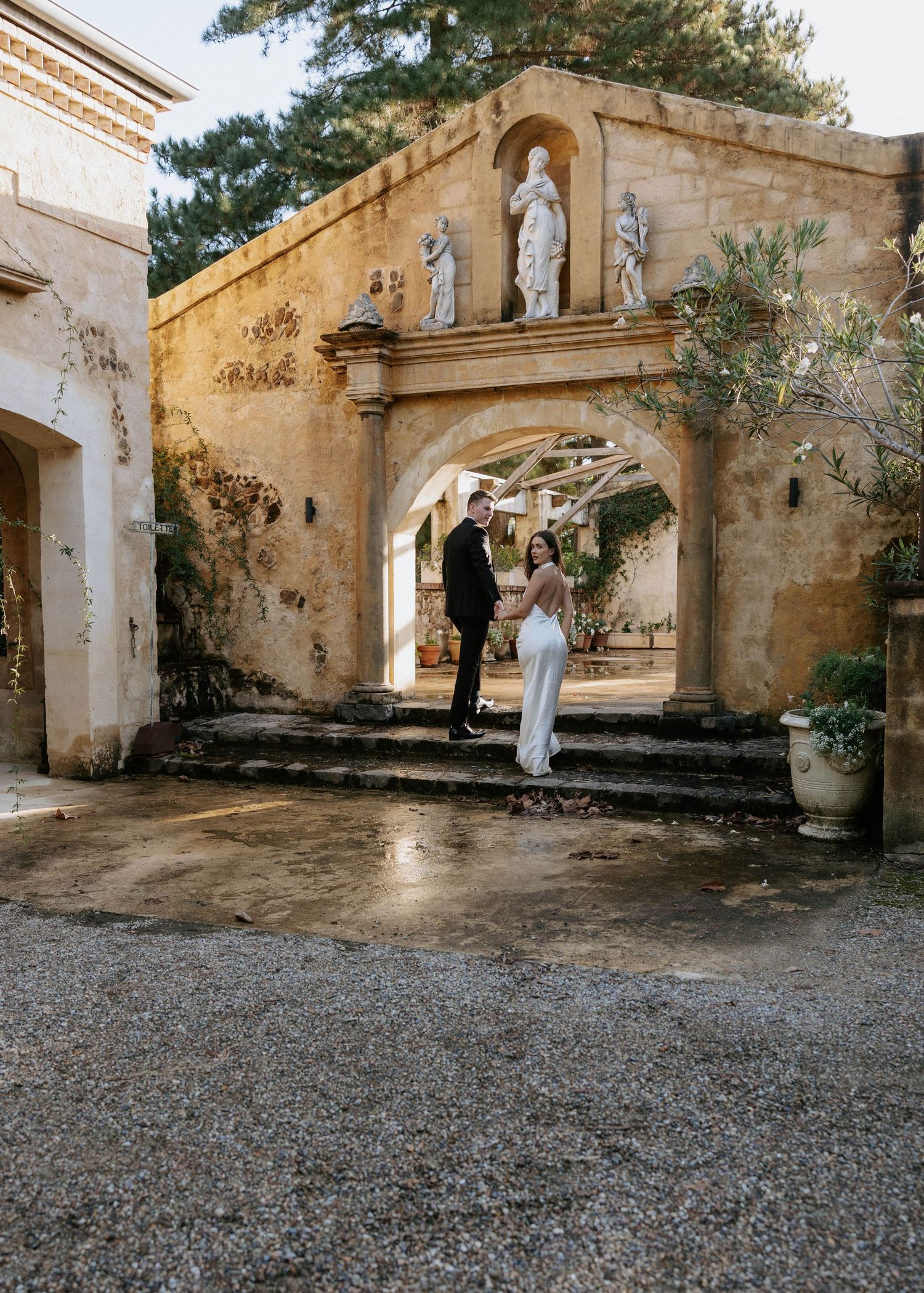 Couple in wedding attire standing before a stone archway with statues at an elegant historic venue.
