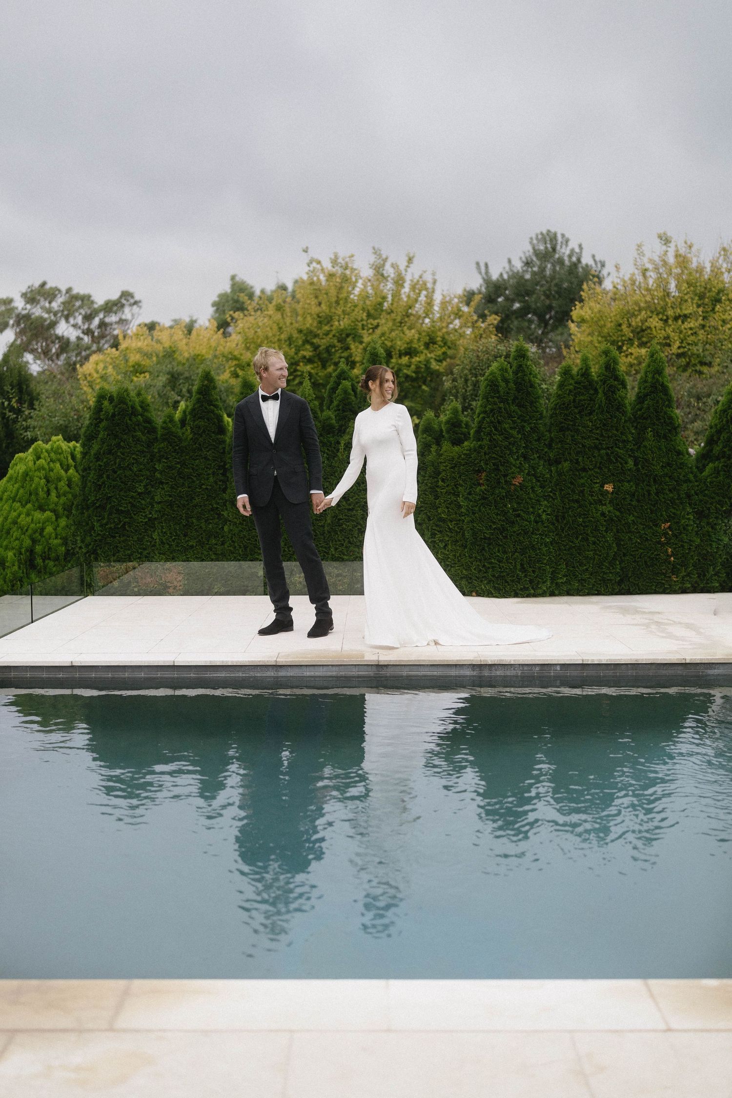 Bride in white gown and groom in tuxedo hold hands beside elegant pool with manicured hedges backdrop.