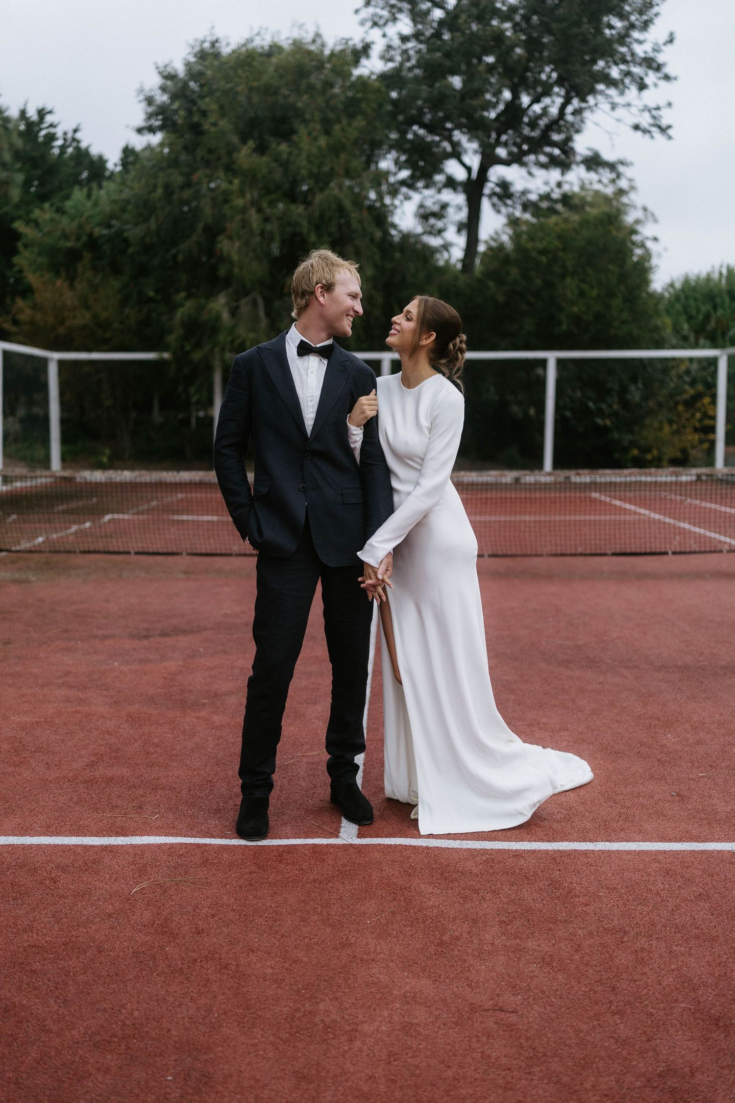 Bride and groom share a romantic moment on a red tennis court, she in a white gown, he in a black tuxedo.