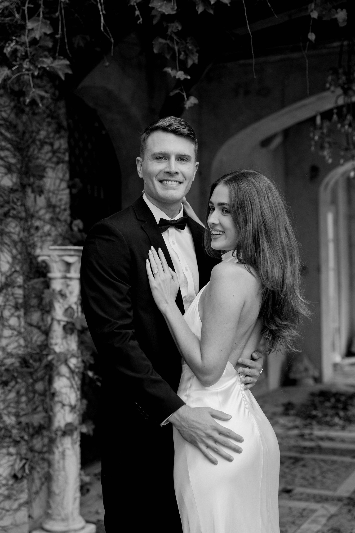 Elegant couple in black and white, man in tuxedo and woman in white gown, posing at a vine-covered estate.