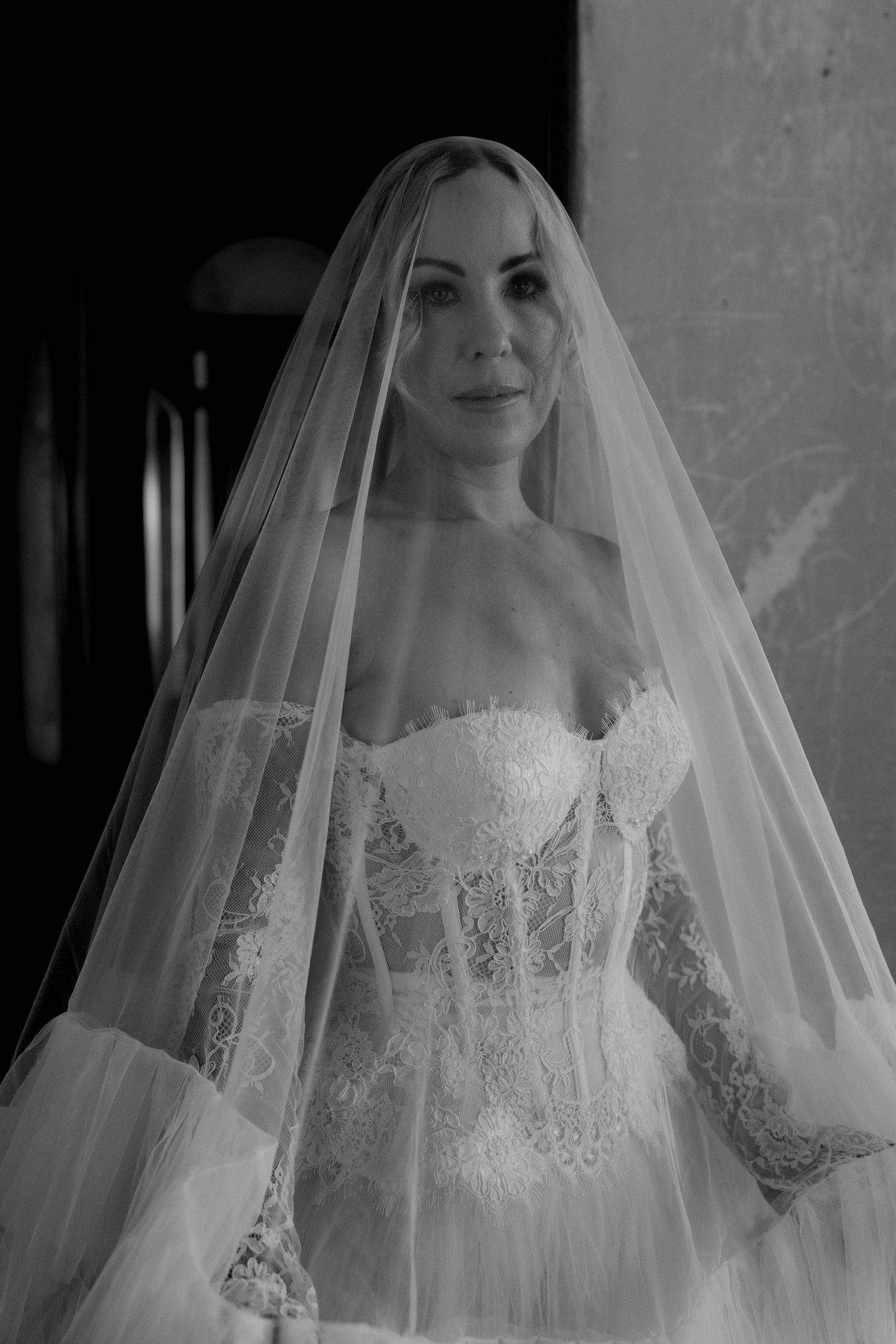 A black and white photograph of a bride wearing a lace corset wedding dress with a long flowing veil.