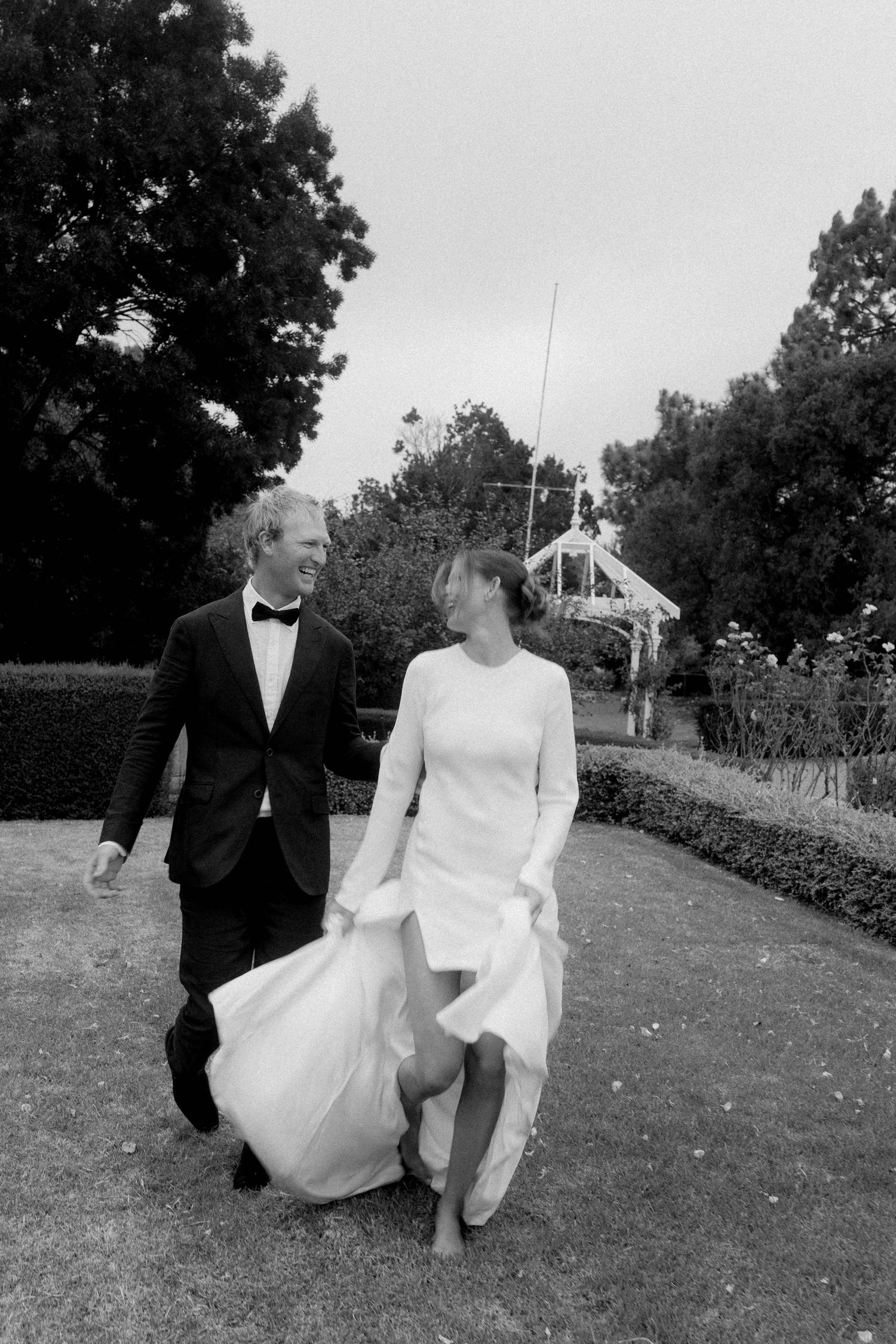 Black and white photo of a couple in wedding attire walking through a garden, smiling at each other.