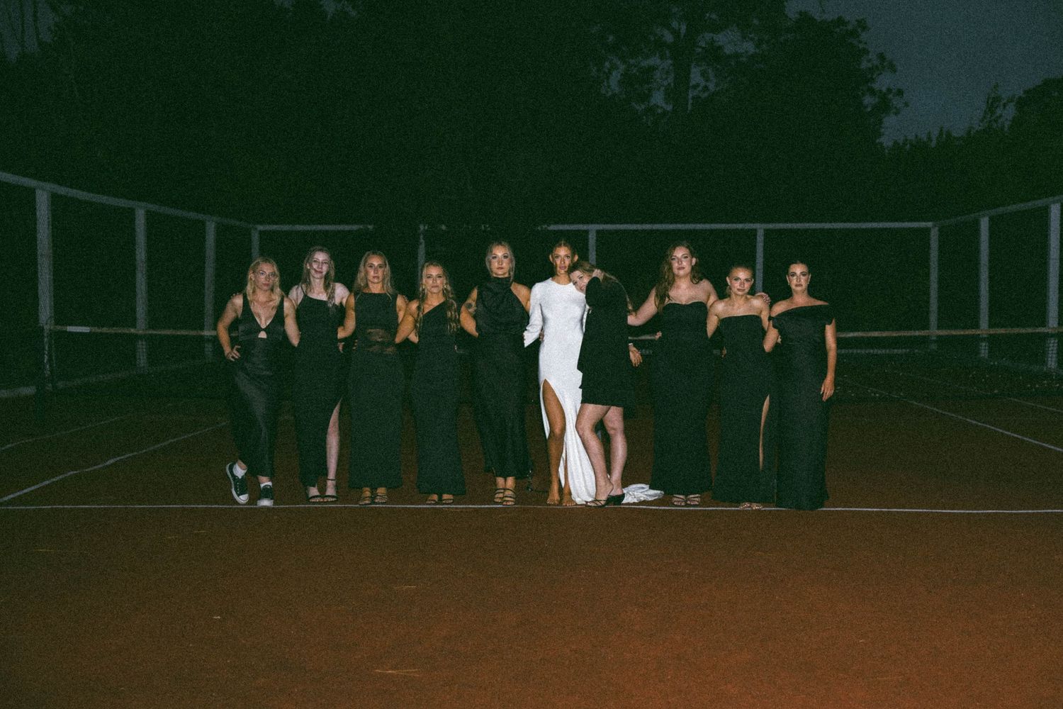 Bride in white gown poses with bridesmaids in black dresses on a tennis court at night.