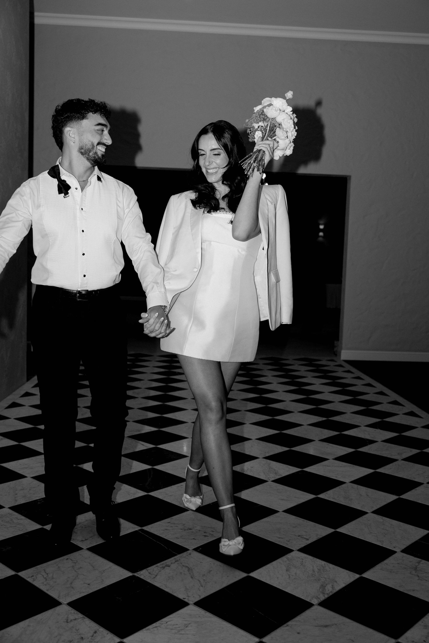 Black and white photo of a smiling bride in a mini dress holding bouquet aloft while walking hand in hand with groom on checkered floor.