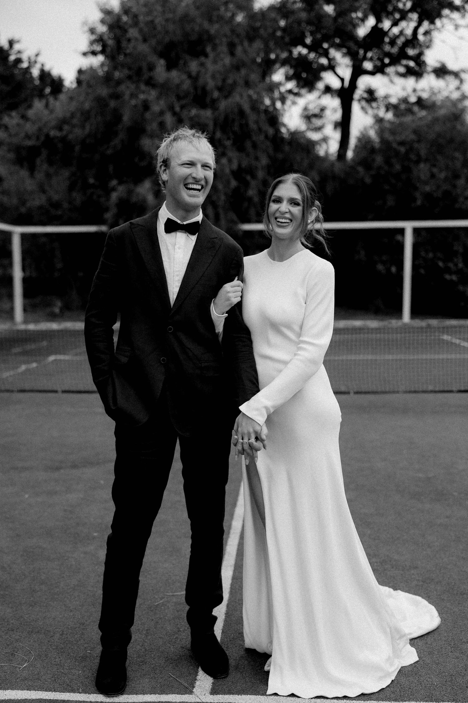 Black and white photo of smiling bride in elegant long-sleeve gown and groom in tuxedo holding hands on a tennis court.