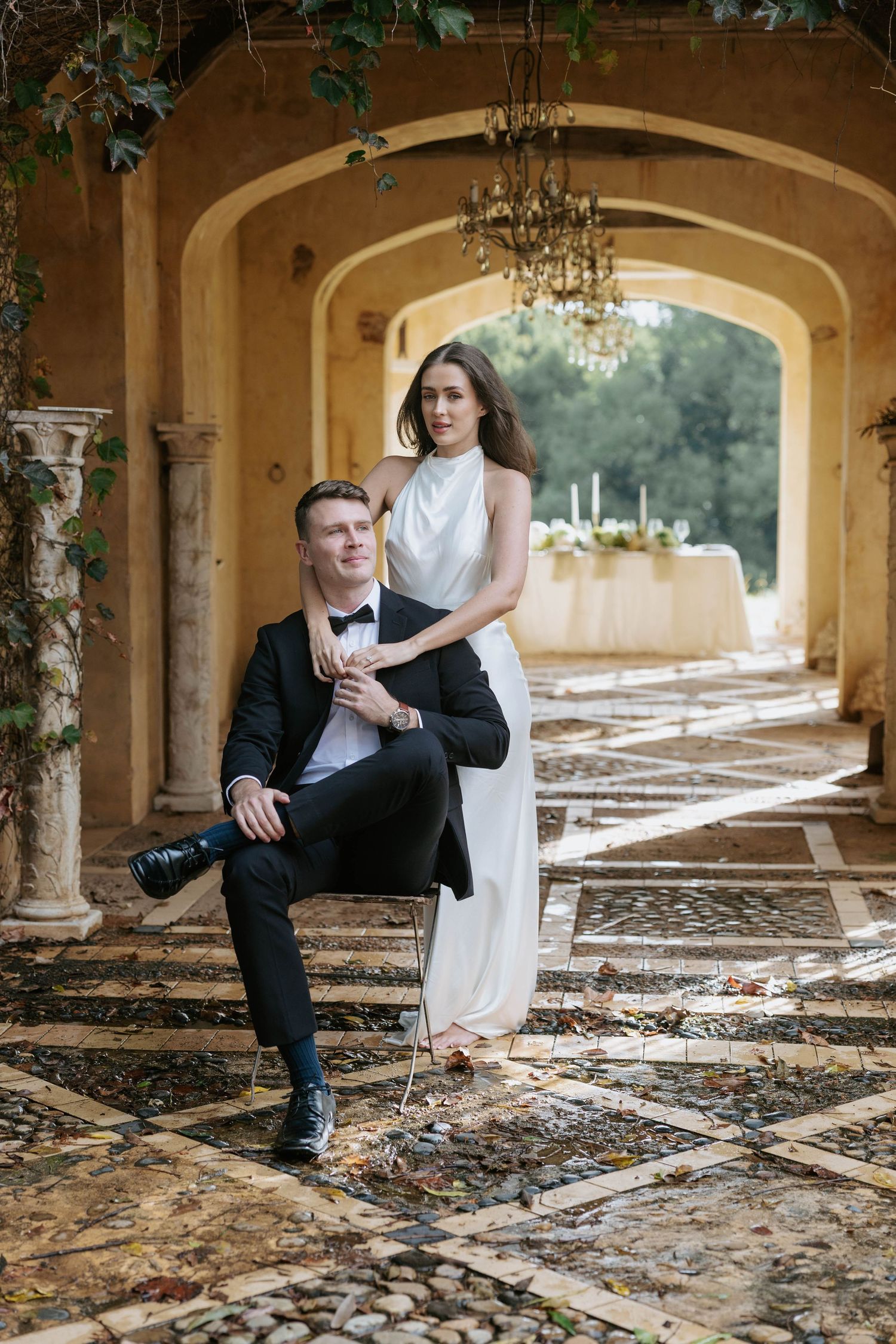 Elegant couple in wedding attire posing under an arched colonnade, groom seated holding camera, bride standing behind him.