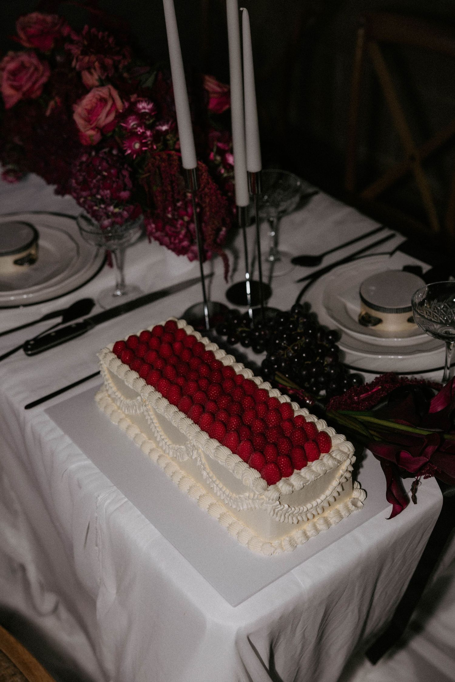 A rectangular cake decorated with red and black berries sits on an elegant table setting with white linens and tall candles.