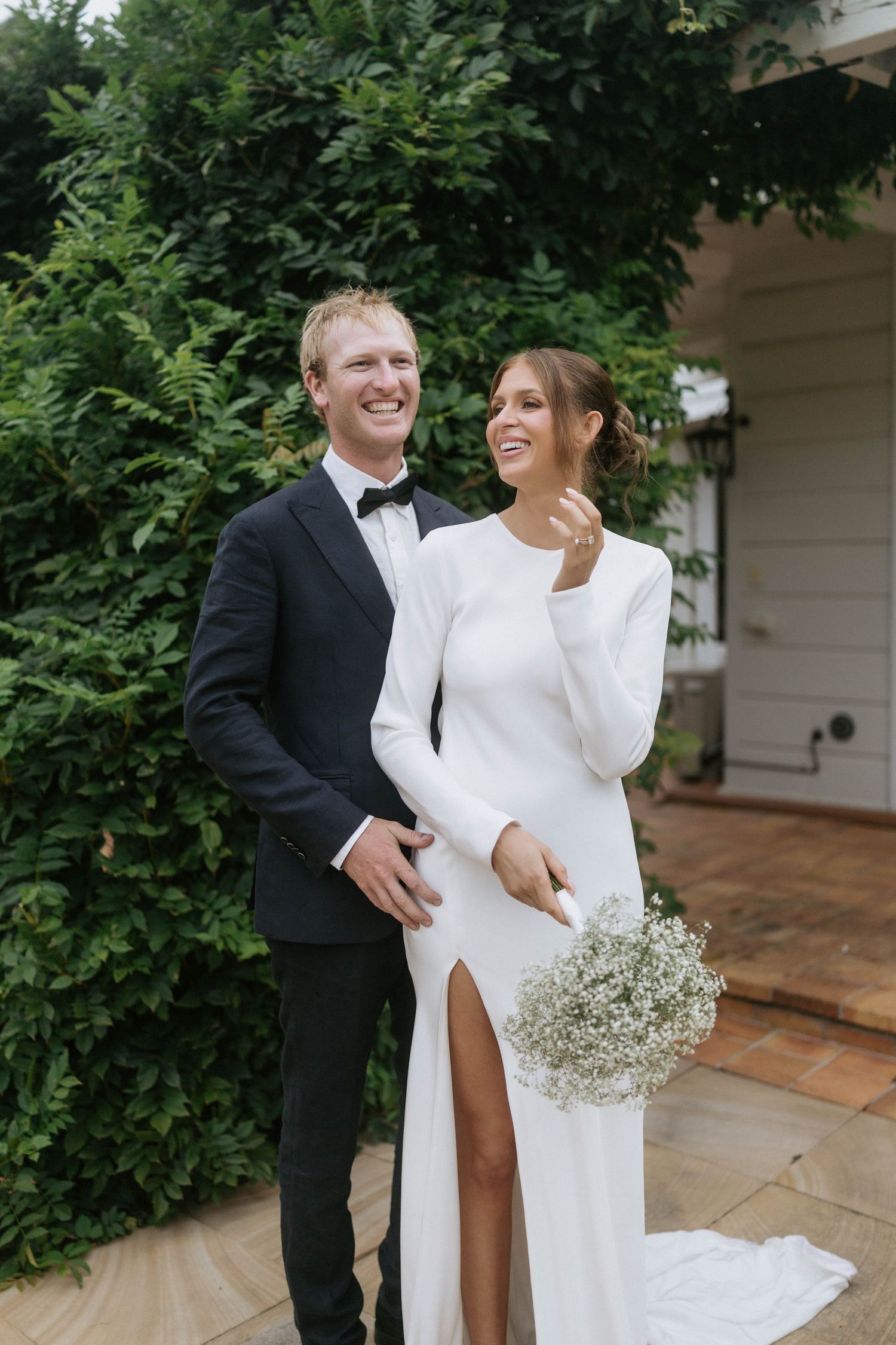 Bride in white long-sleeve gown with slit holds baby's breath bouquet beside groom in black tuxedo outdoors.