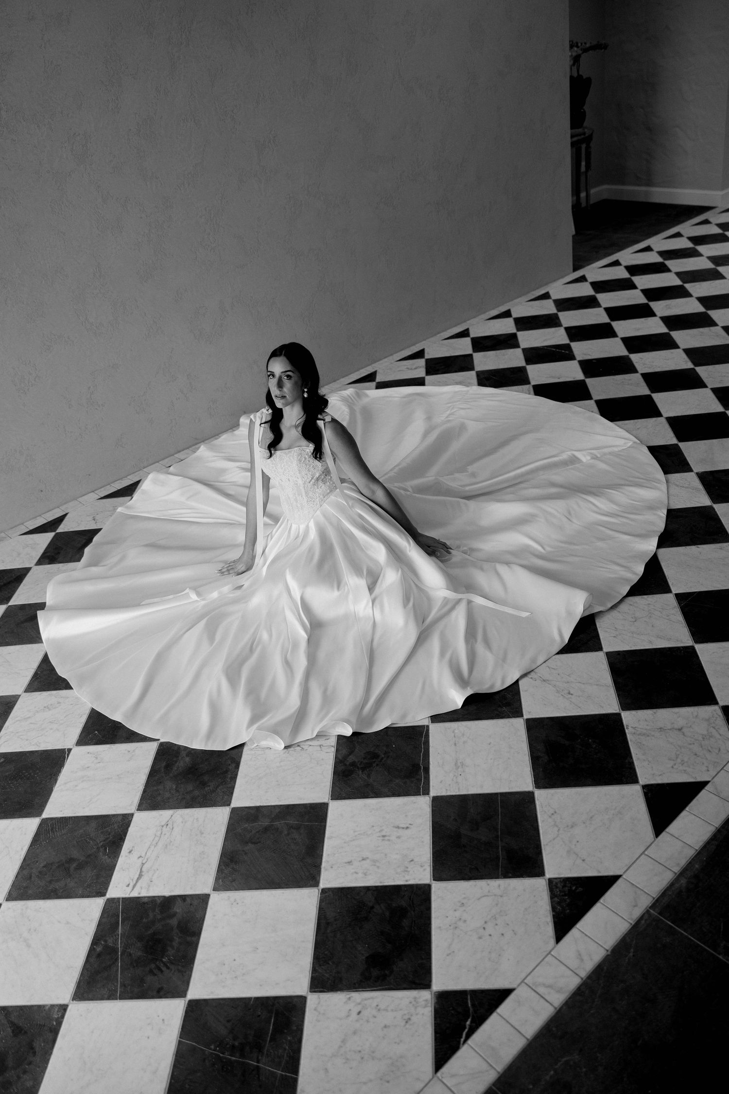 Bride in flowing white gown seated on black and white checkered marble floor, black and white photo.