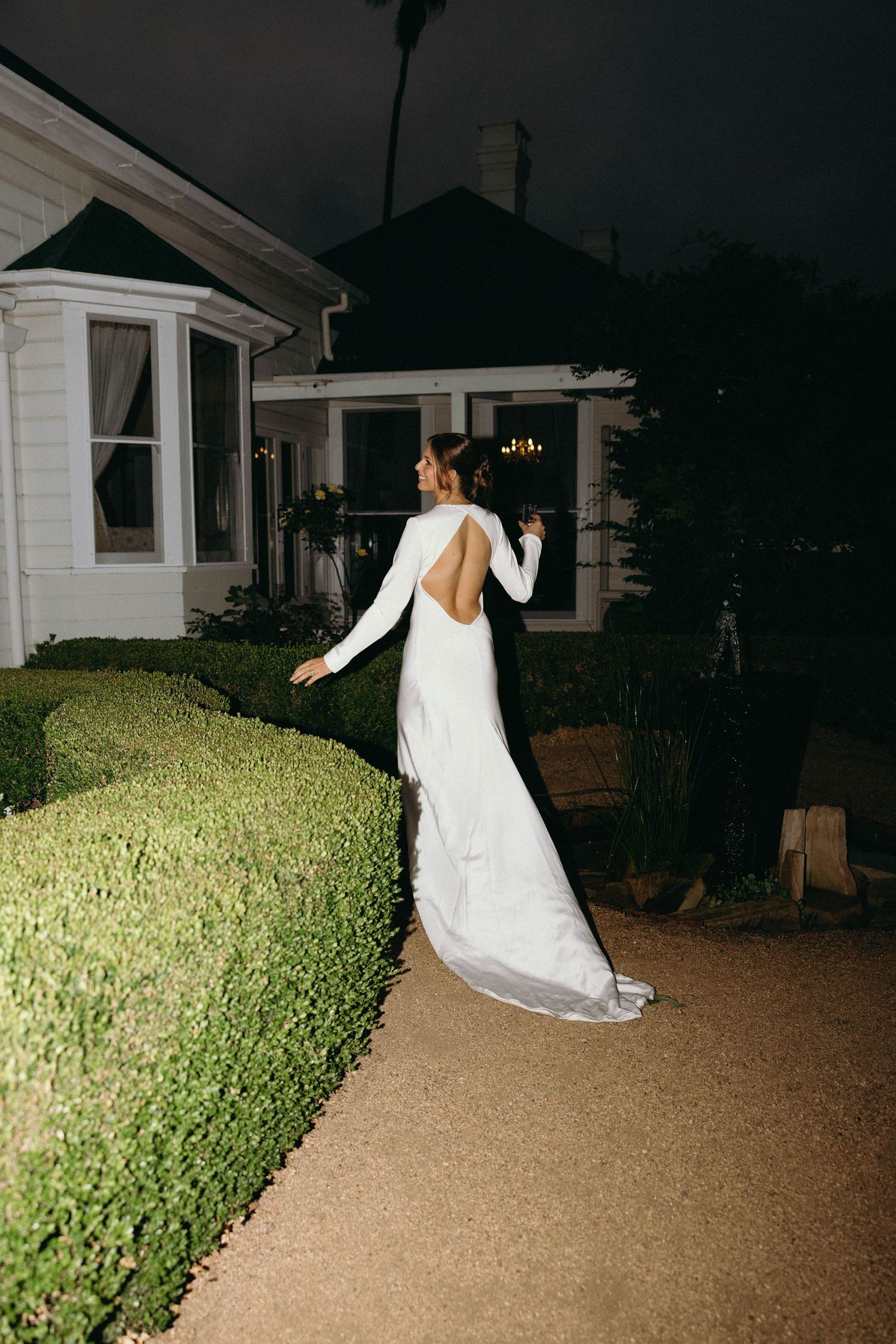Bride in elegant backless white gown with long sleeves posing beside hedges at night.