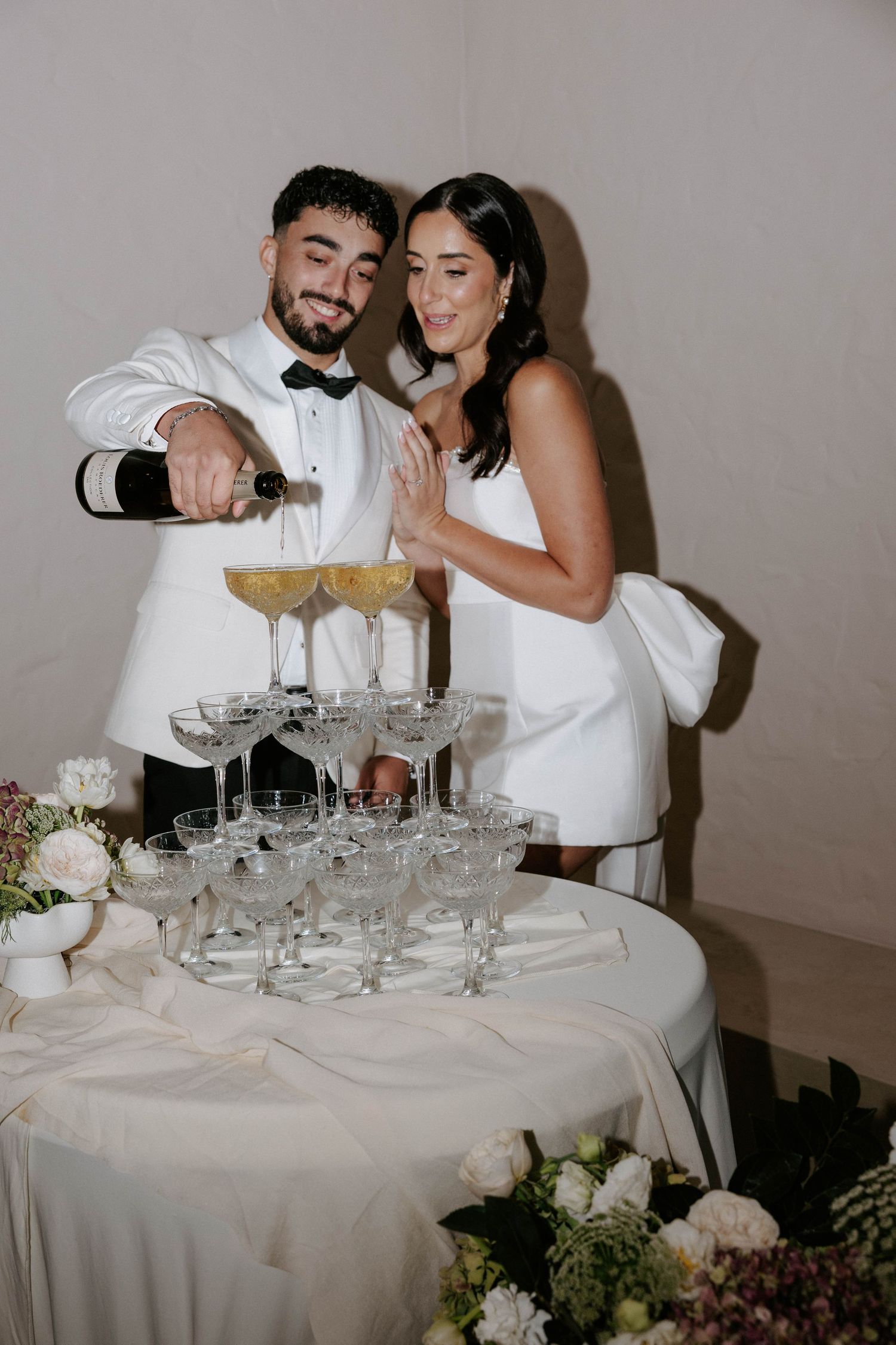 Bride and groom pouring champagne tower at wedding reception, both dressed in white formal attire.