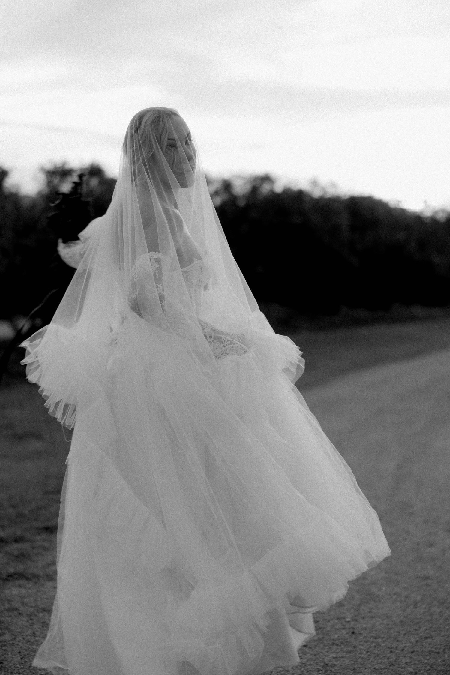 Black and white photo of a bride in a flowing tulle wedding gown and veil, walking outdoors at dusk.