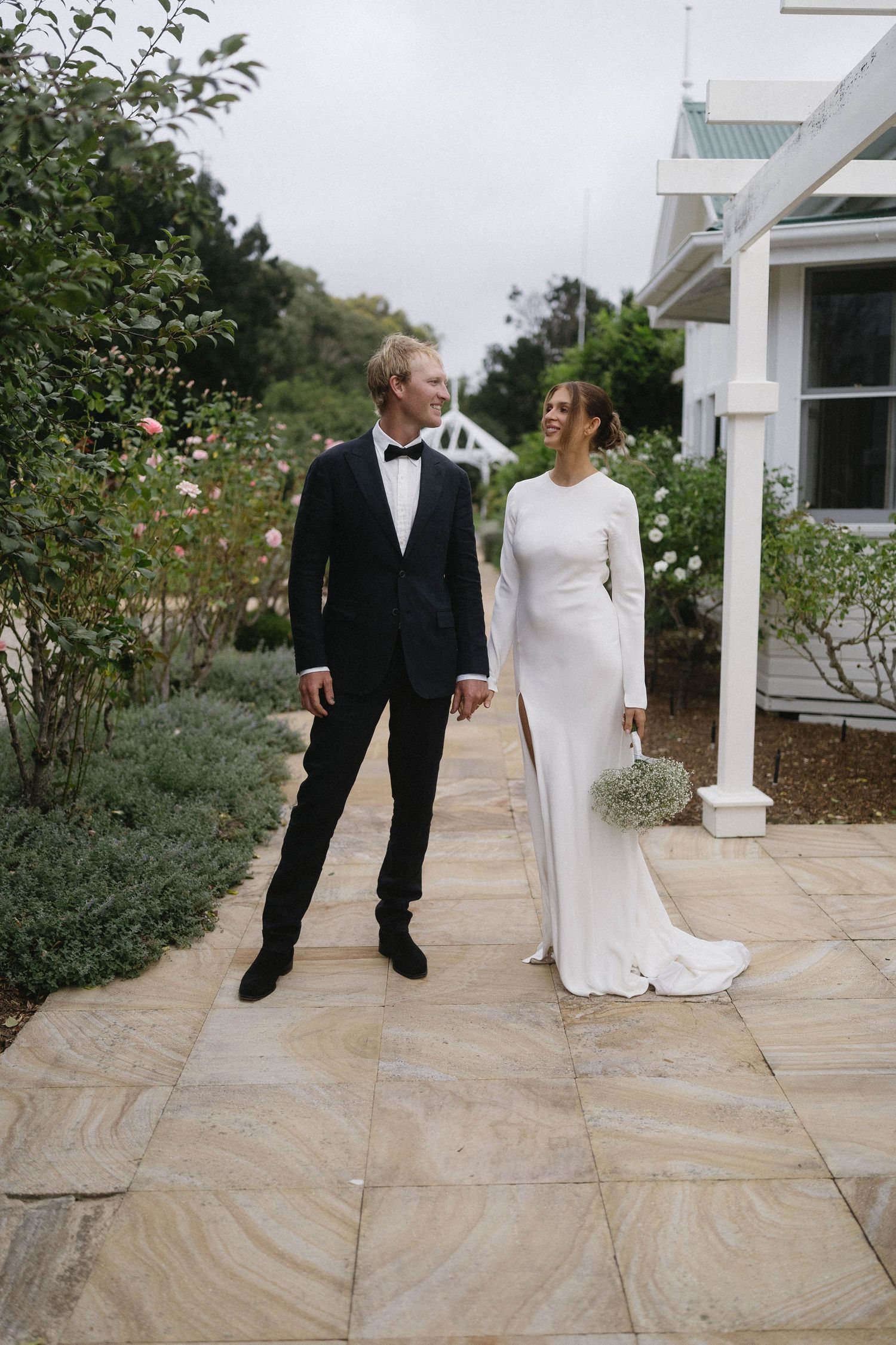Bride and groom walk hand in hand along a stone garden path, she in a sleek white gown, he in a classic black tuxedo.