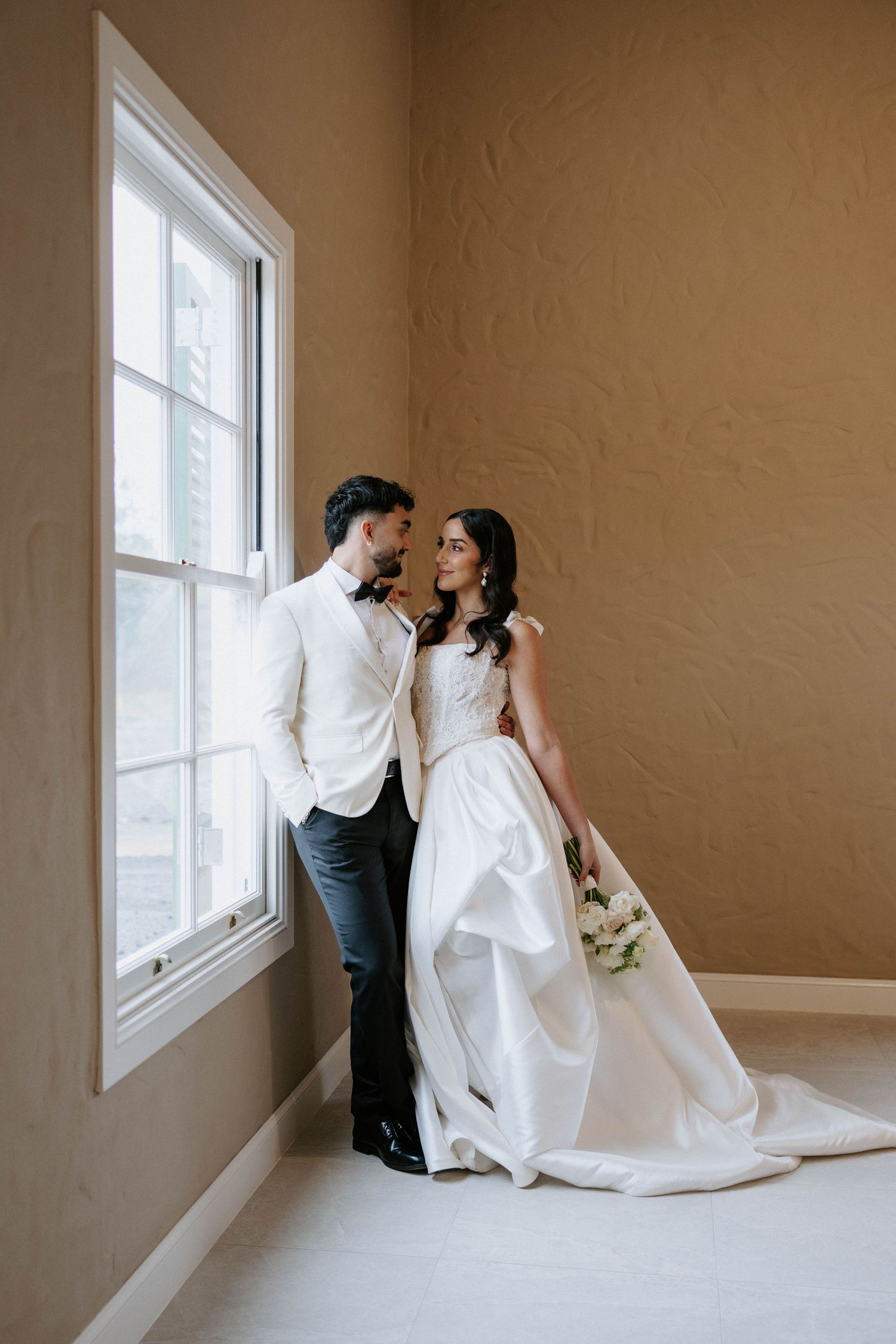 Bride and groom sharing romantic moment by window, bride holding white bouquet in elegant satin wedding gown.
