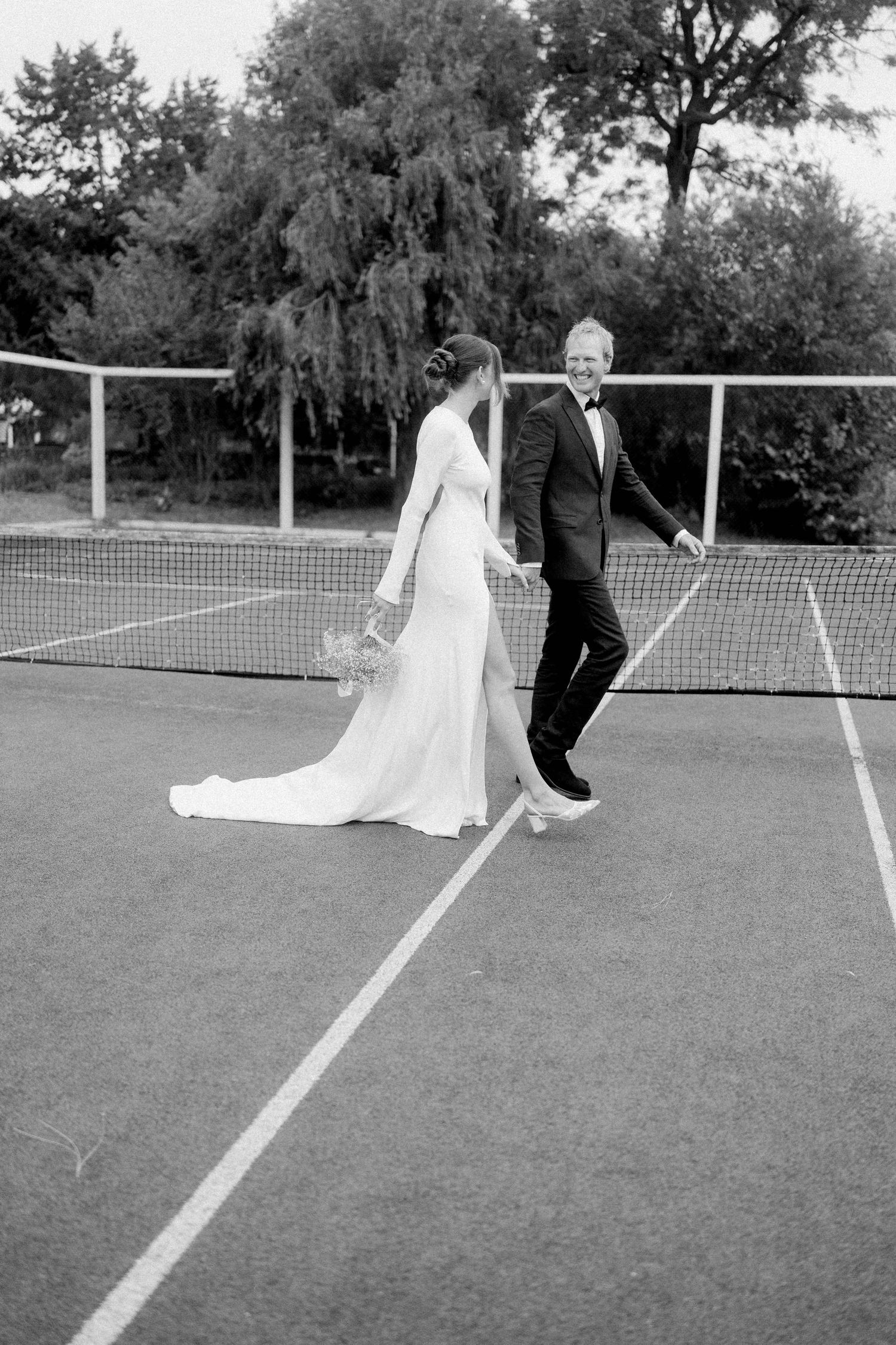 Black and white photo of bride and groom in formal wear walking hand in hand on tennis court.