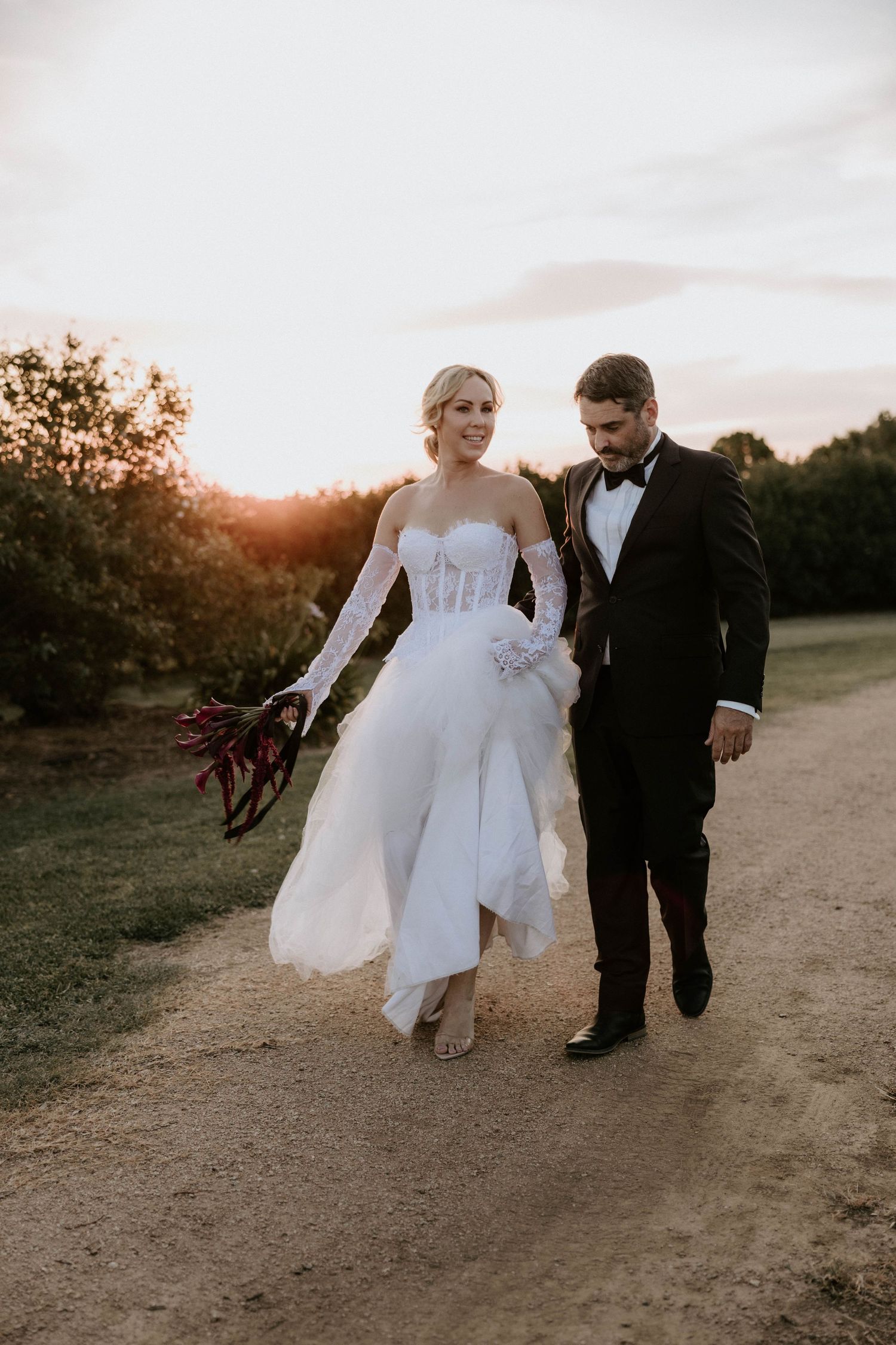 A couple in formal wedding attire walks together on a dirt path at sunset while holding hands.
