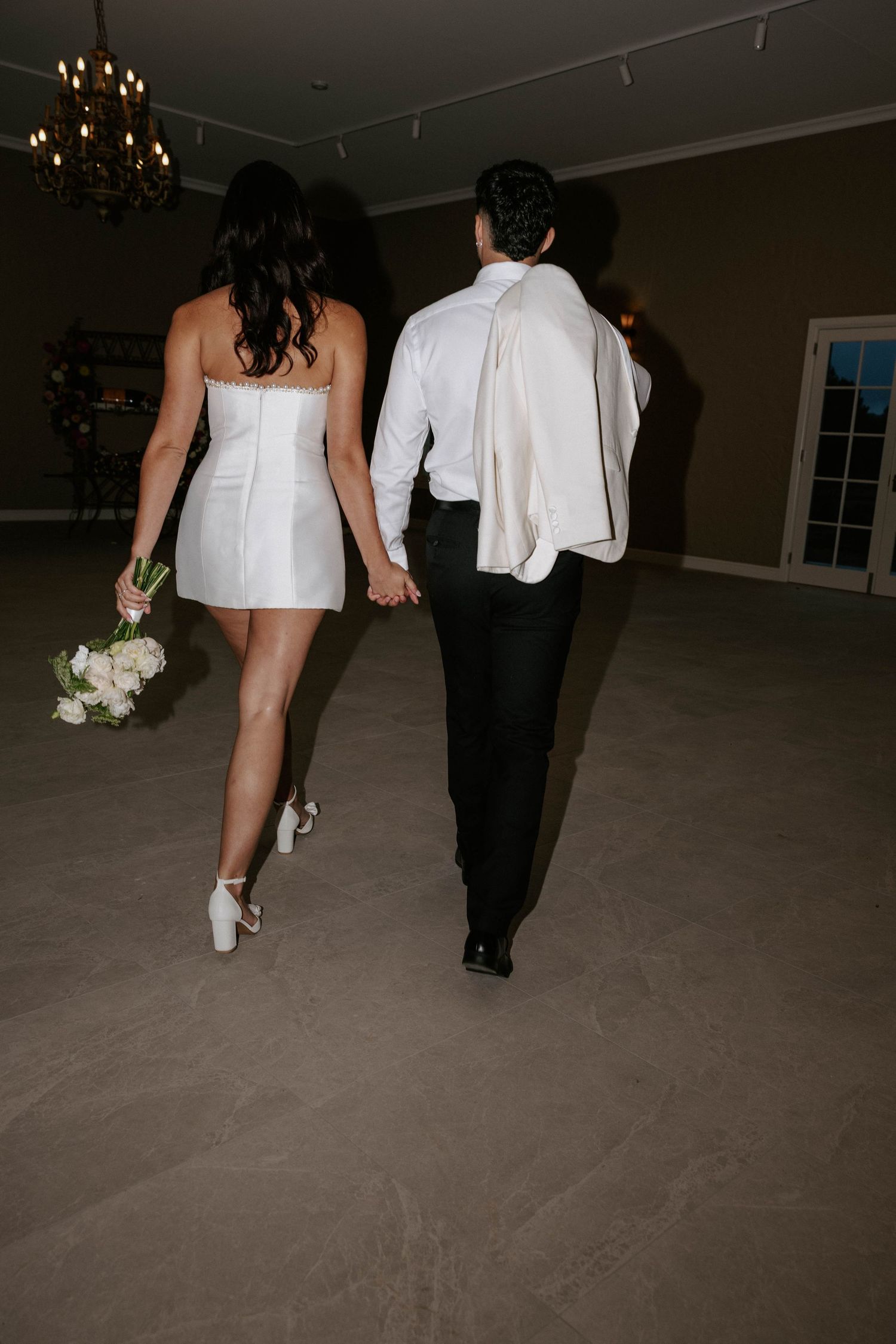 Bride in short white mini dress and groom in black tuxedo walking hand in hand across ballroom floor.