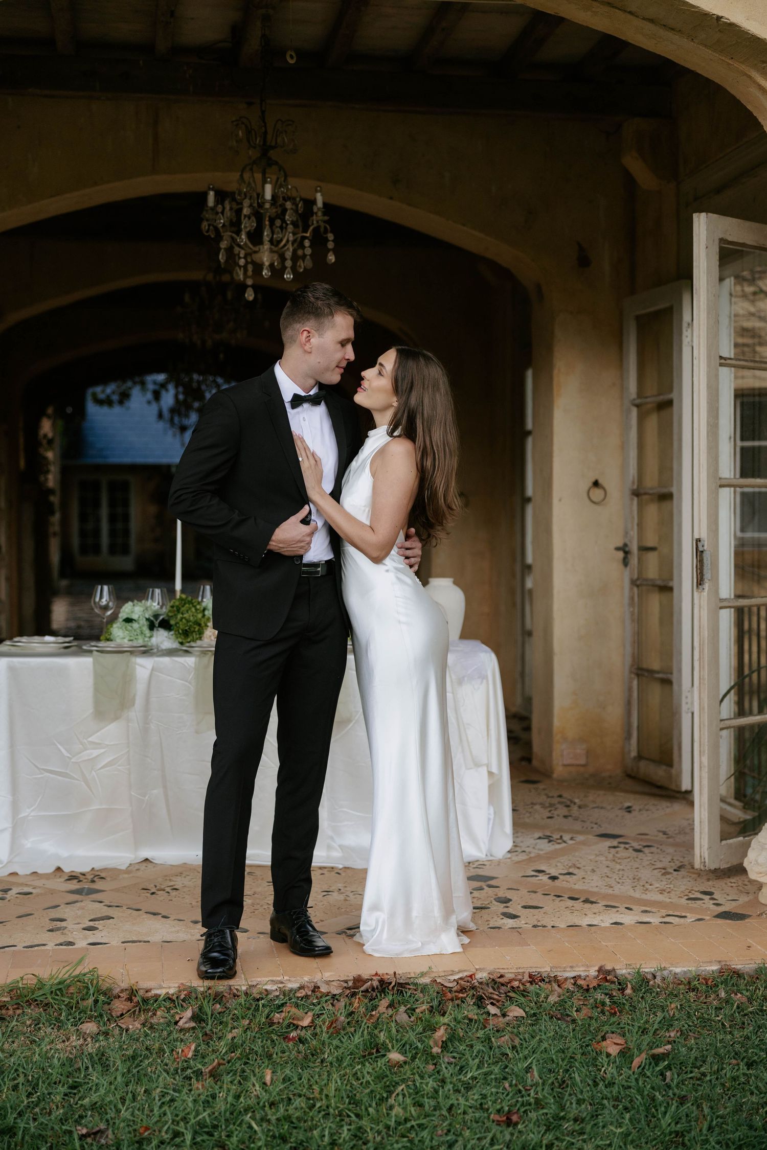 Elegant bride and groom sharing a romantic moment outside a rustic villa, bride in white gown, groom in black tuxedo.