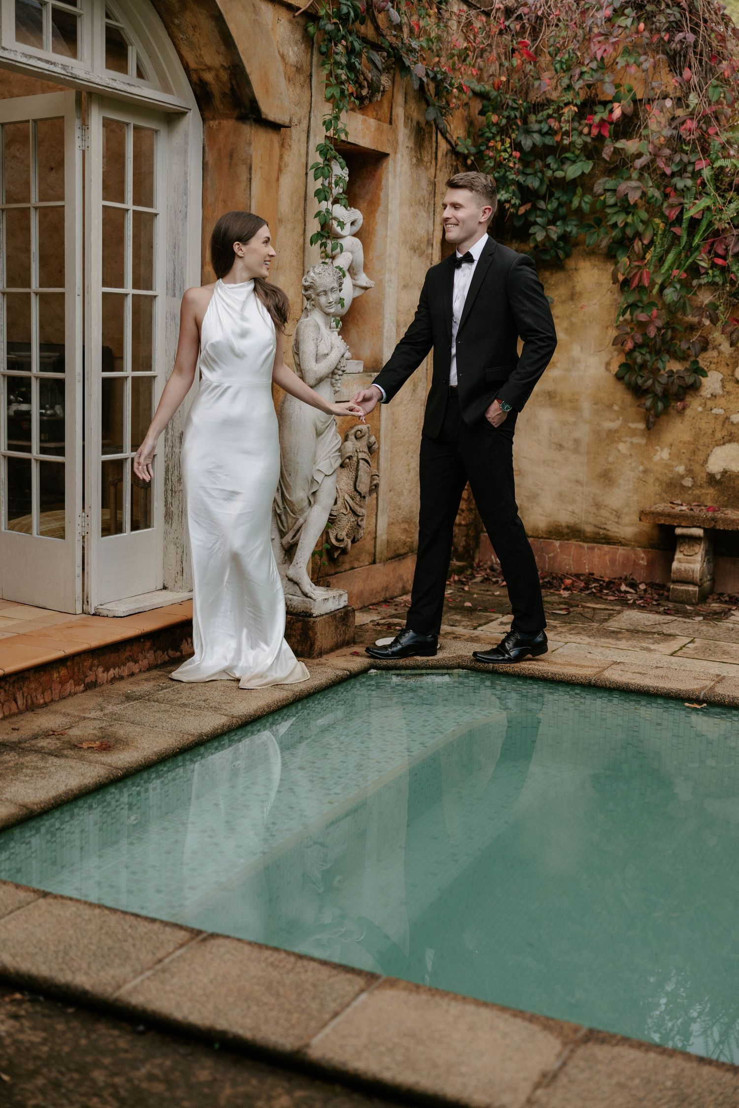 Bride in white halter gown and groom in black tuxedo hold hands beside a garden pool at an elegant venue.