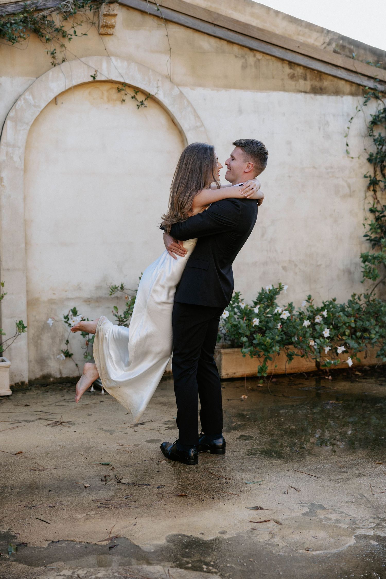 Groom lifting bride in white dress outdoors against rustic stone wall with greenery at elegant wedding venue.