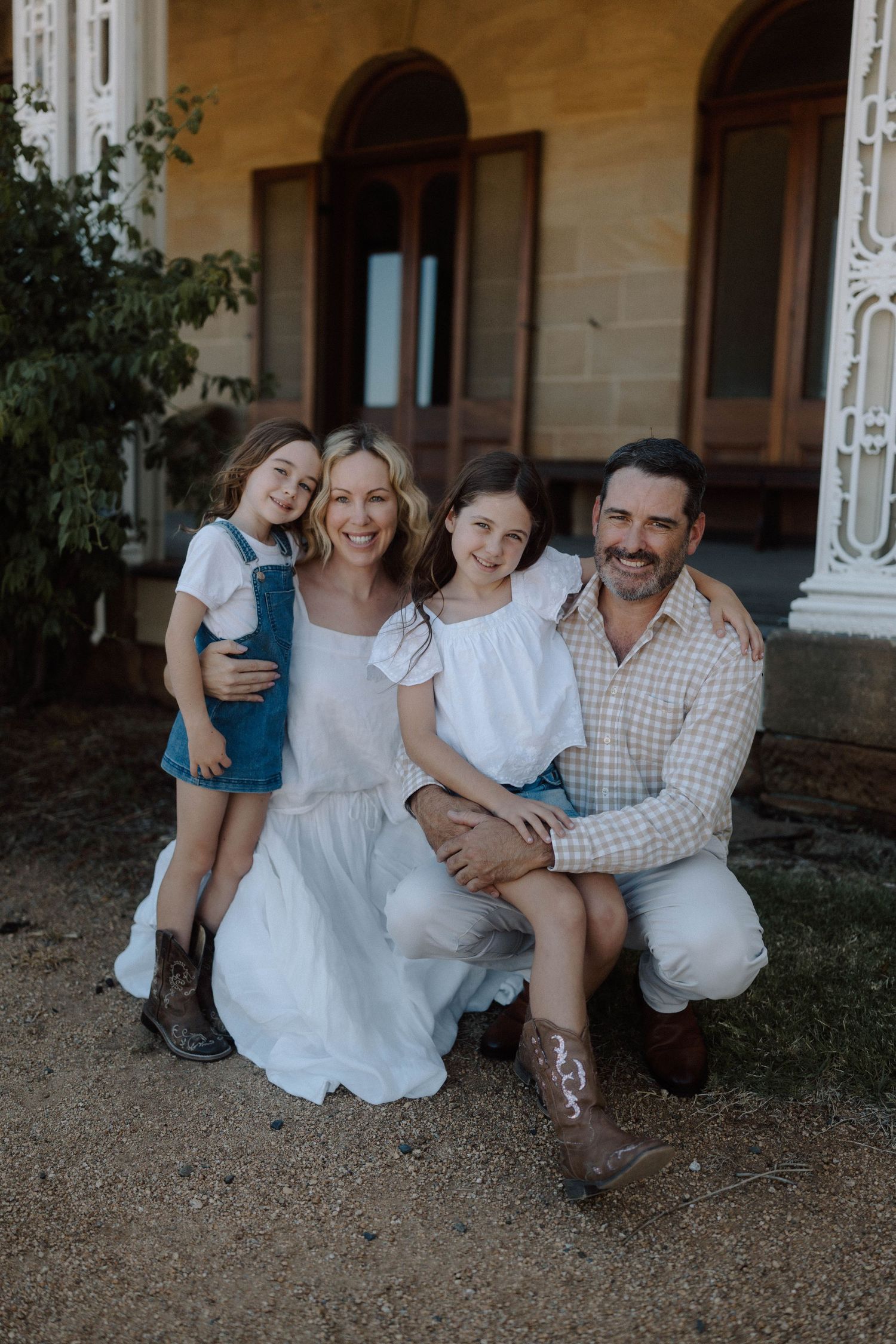A smiling family of four poses outdoors in front of a historic building, with two young girls and their parents.