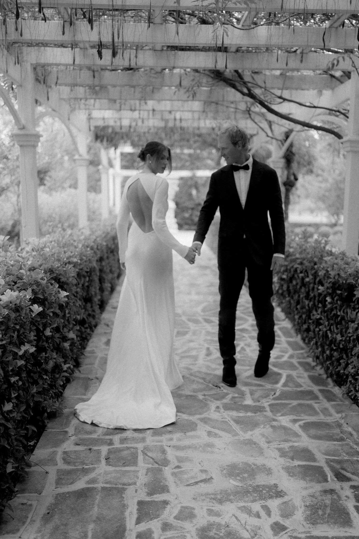 Black and white photo of bride and groom holding hands walking under pergola.
