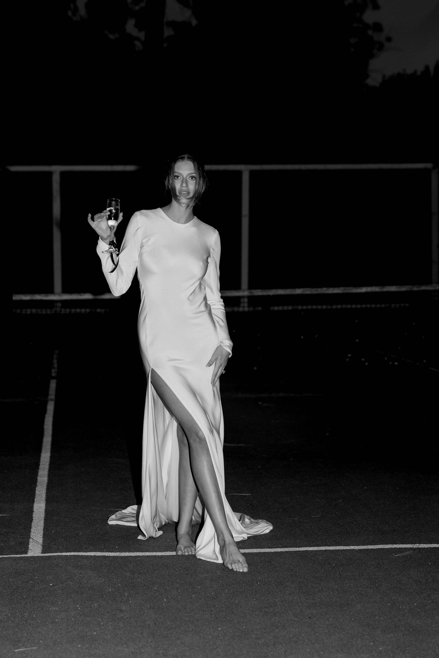 Black and white photo of a woman in an elegant white gown holding a champagne glass on a tennis court at night.