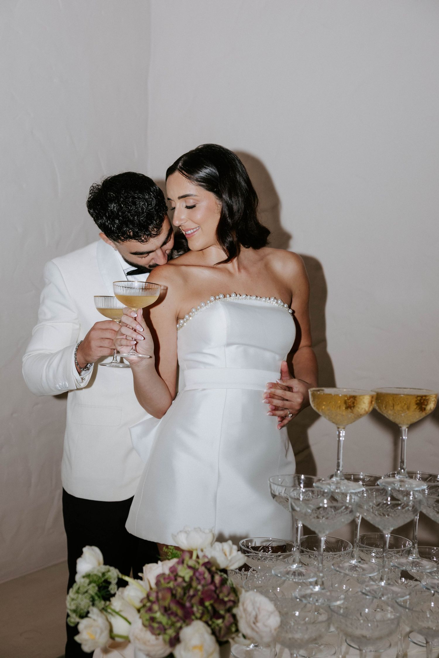 Bride and groom sharing champagne coupe glasses at wedding reception tower display in elegant white attire.