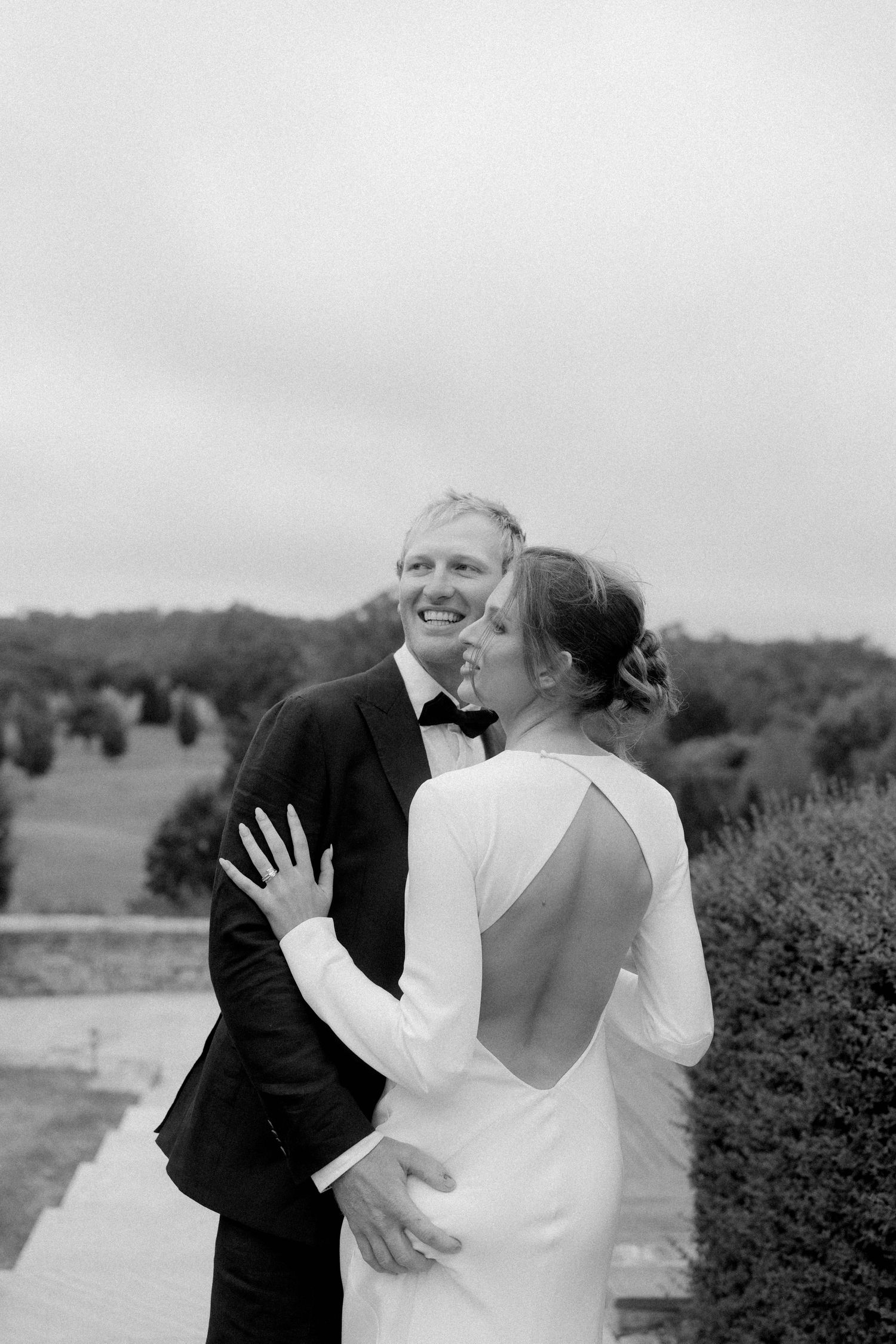 Black and white photo of a smiling groom in a tuxedo embracing his bride in a backless white gown outdoors.