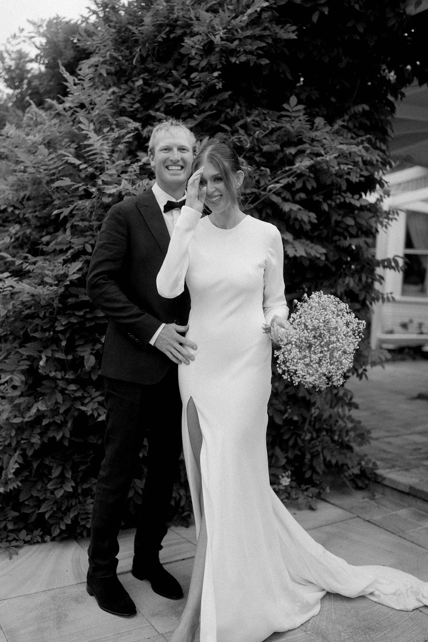 Black and white photo of smiling bride in elegant white gown holding baby's breath bouquet with groom in tuxedo outdoors.
