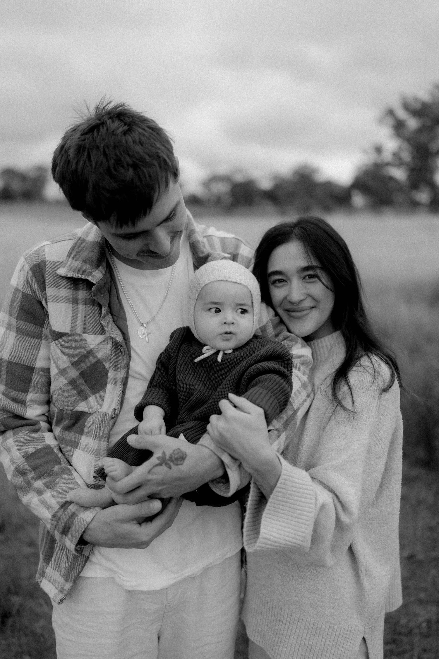 Black and white photo of young parents smiling and holding their baby outdoors in a field.