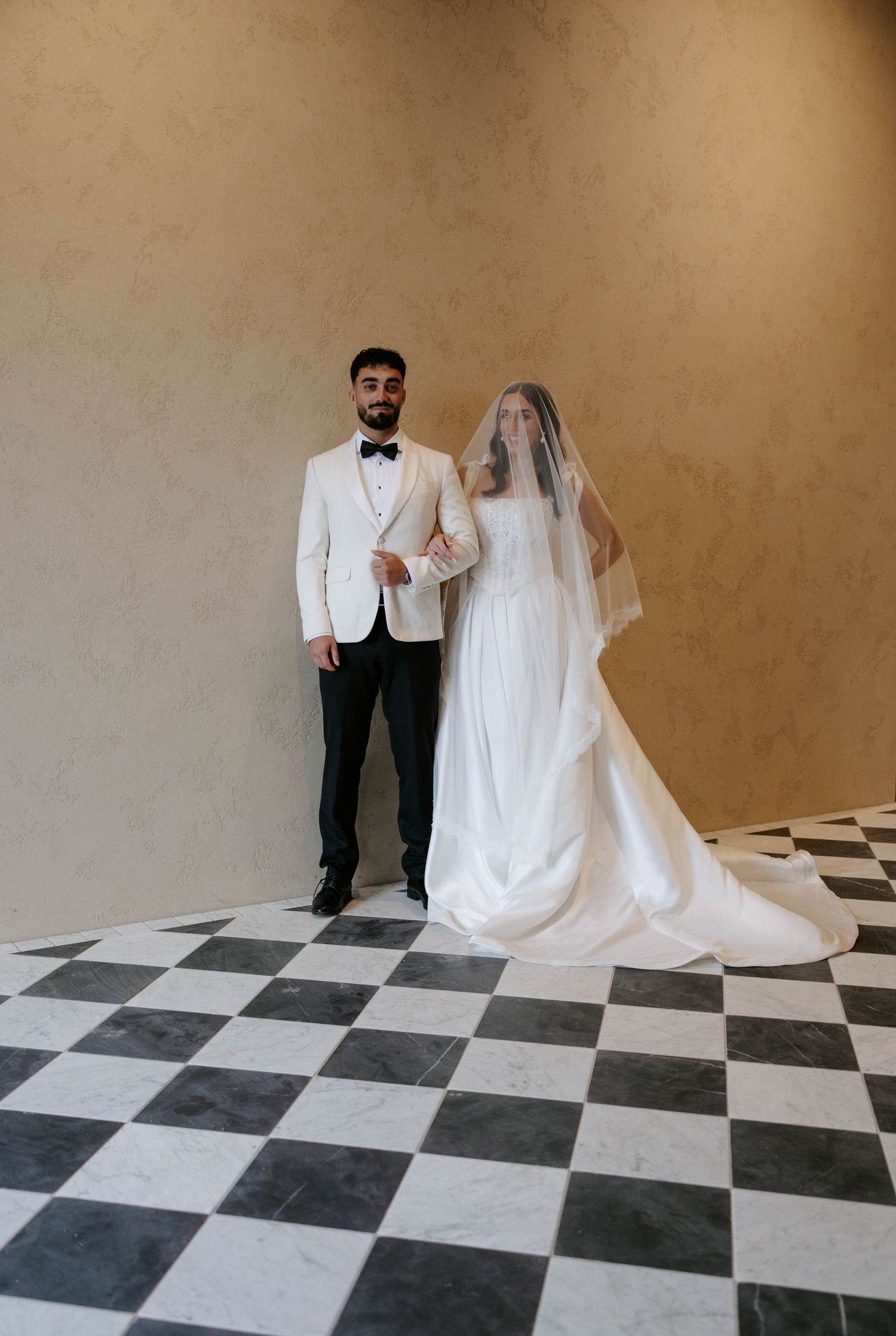 Bride and groom posing on checkered marble floor, groom in white tuxedo jacket, bride in flowing white gown with veil.
