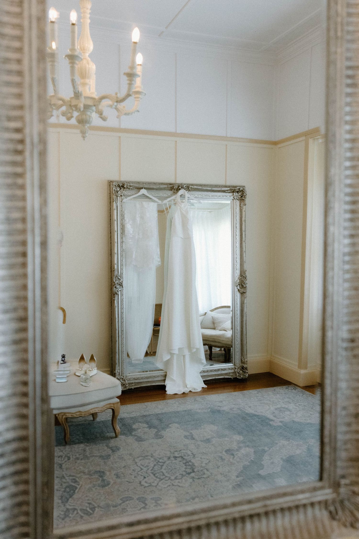 An elegant white wedding dress hangs on display in front of an ornate silver mirror with a chandelie. gabbinbar homestead toowoomba
