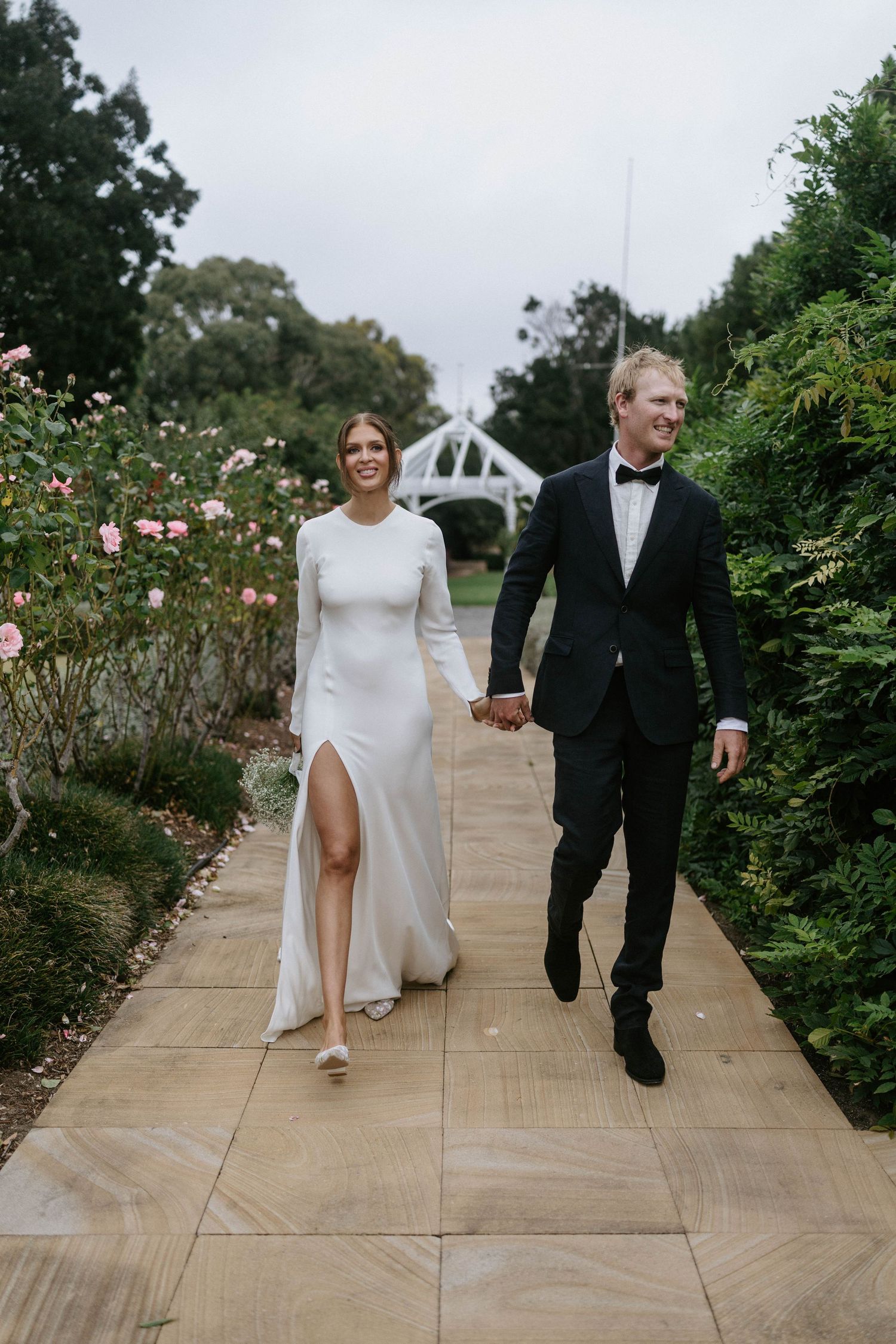 Smiling bride and groom walking hand in hand through a rose garden, bride in sleek white gown with slit.