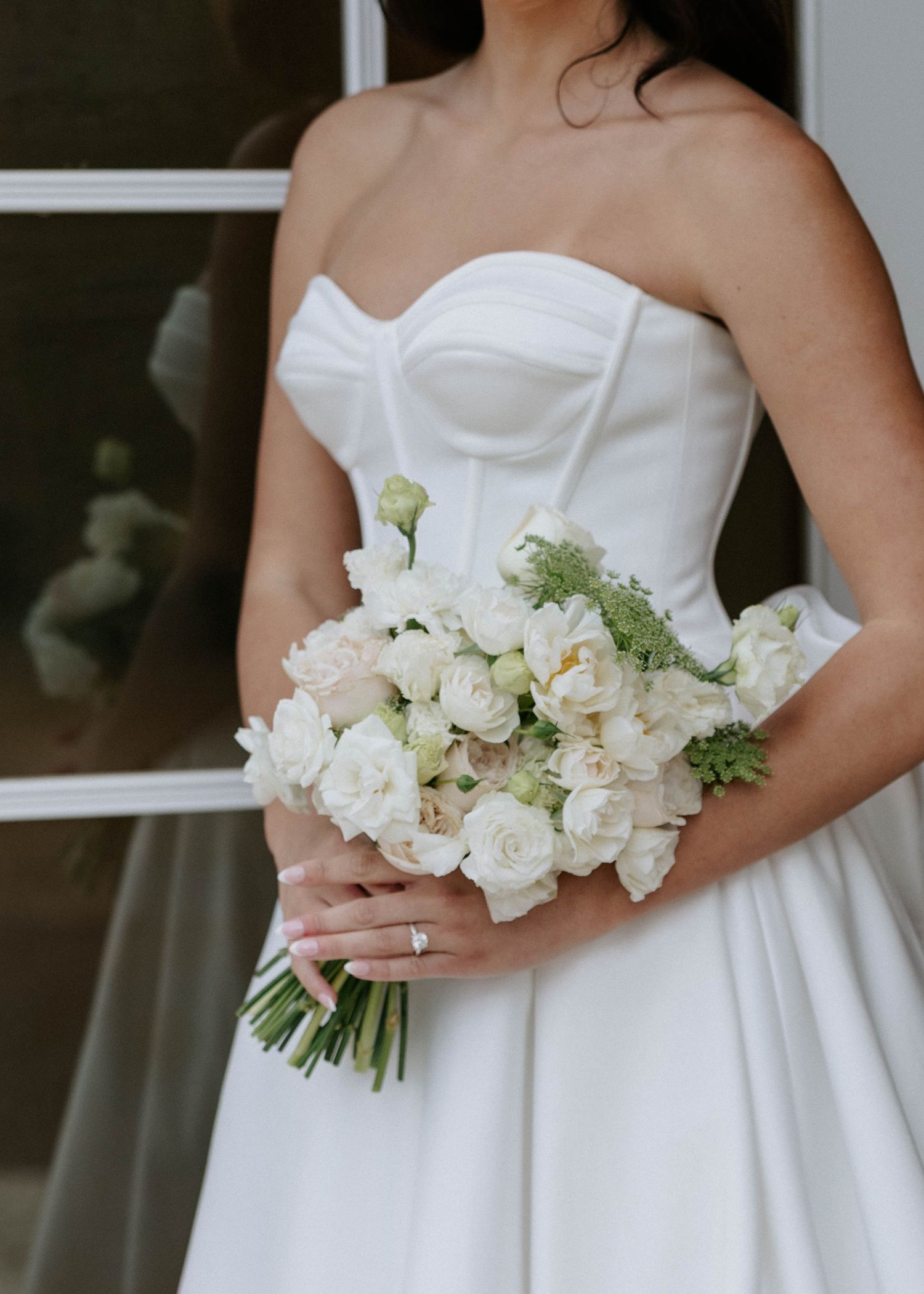 Bride in strapless white satin gown holds lush bouquet of white roses and peonies on wedding day.