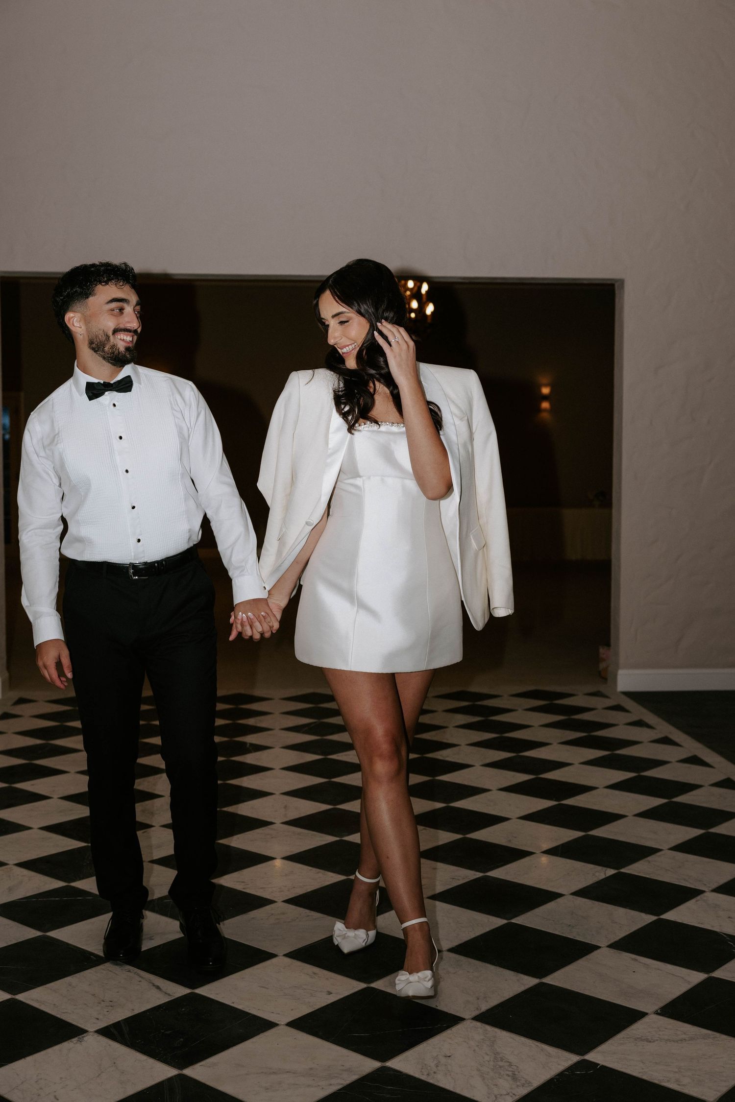 Couple holding hands on checkered floor, groom in tuxedo, bride in mini dress with white blazer.