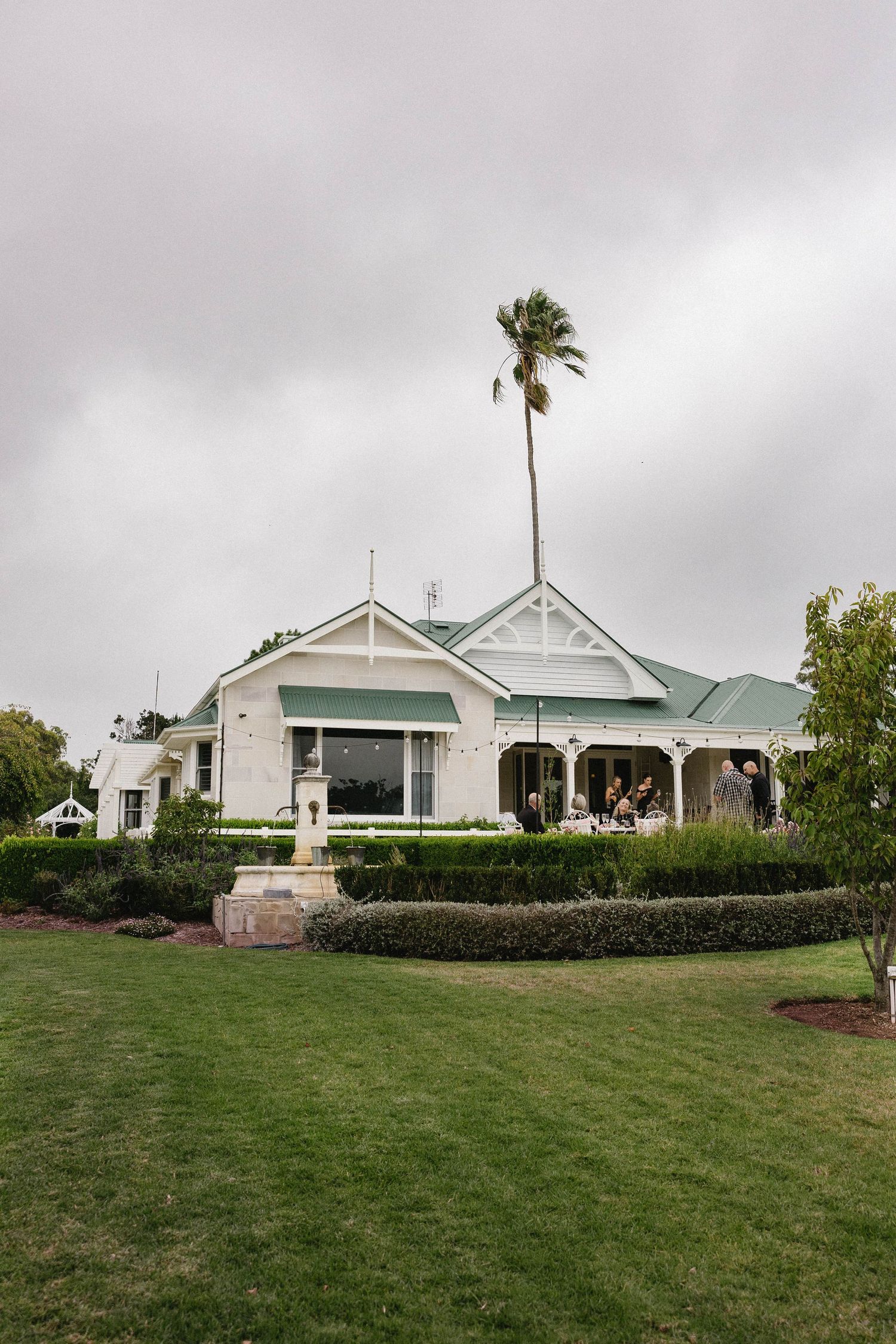 Elegant white Victorian-style wedding venue with green roof and palm tree on overcast day.