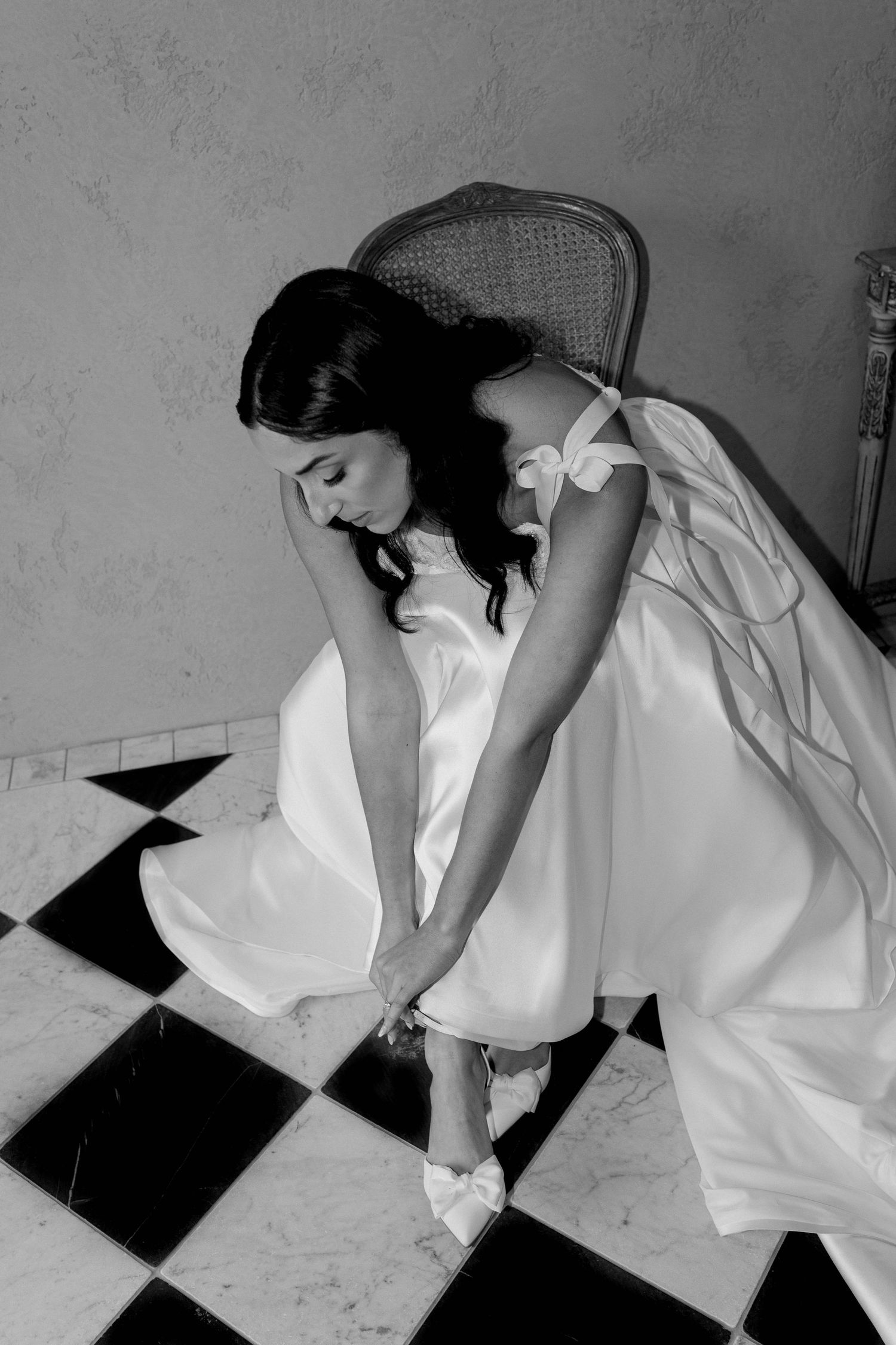 Bride in white wedding dress adjusting her bow heels on black and white checkered floor in black and white photo.