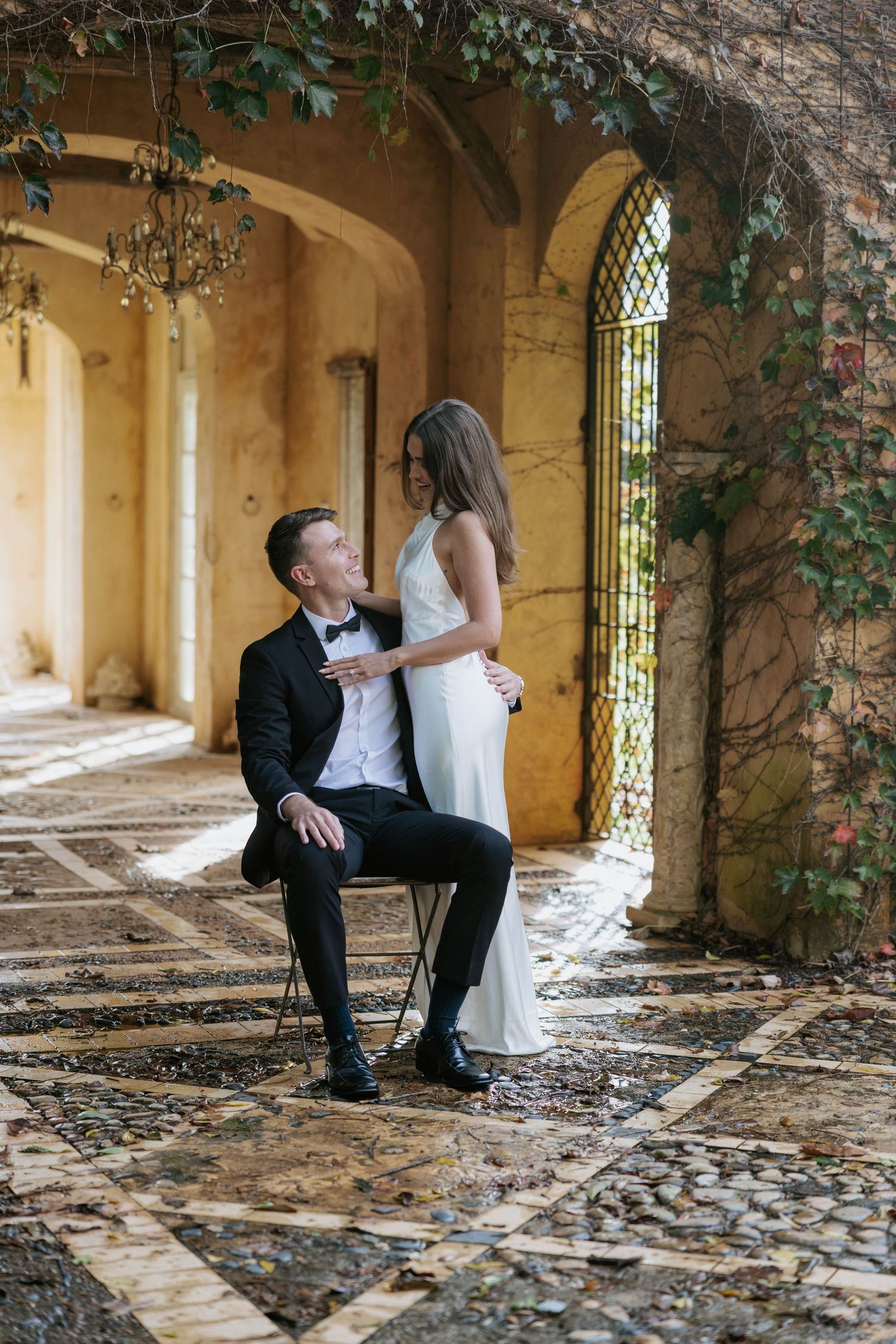 Bride and groom posing in a rustic vine-covered archway, groom seated on chair with bride standing close.