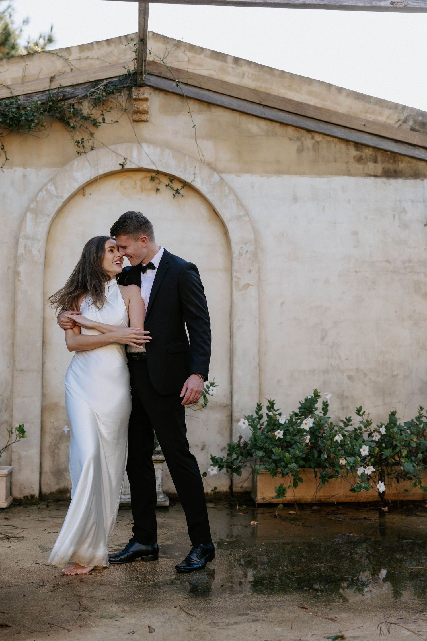 Bride in a sleek white gown and groom in a black tuxedo embracing in front of a rustic arched stone wall.