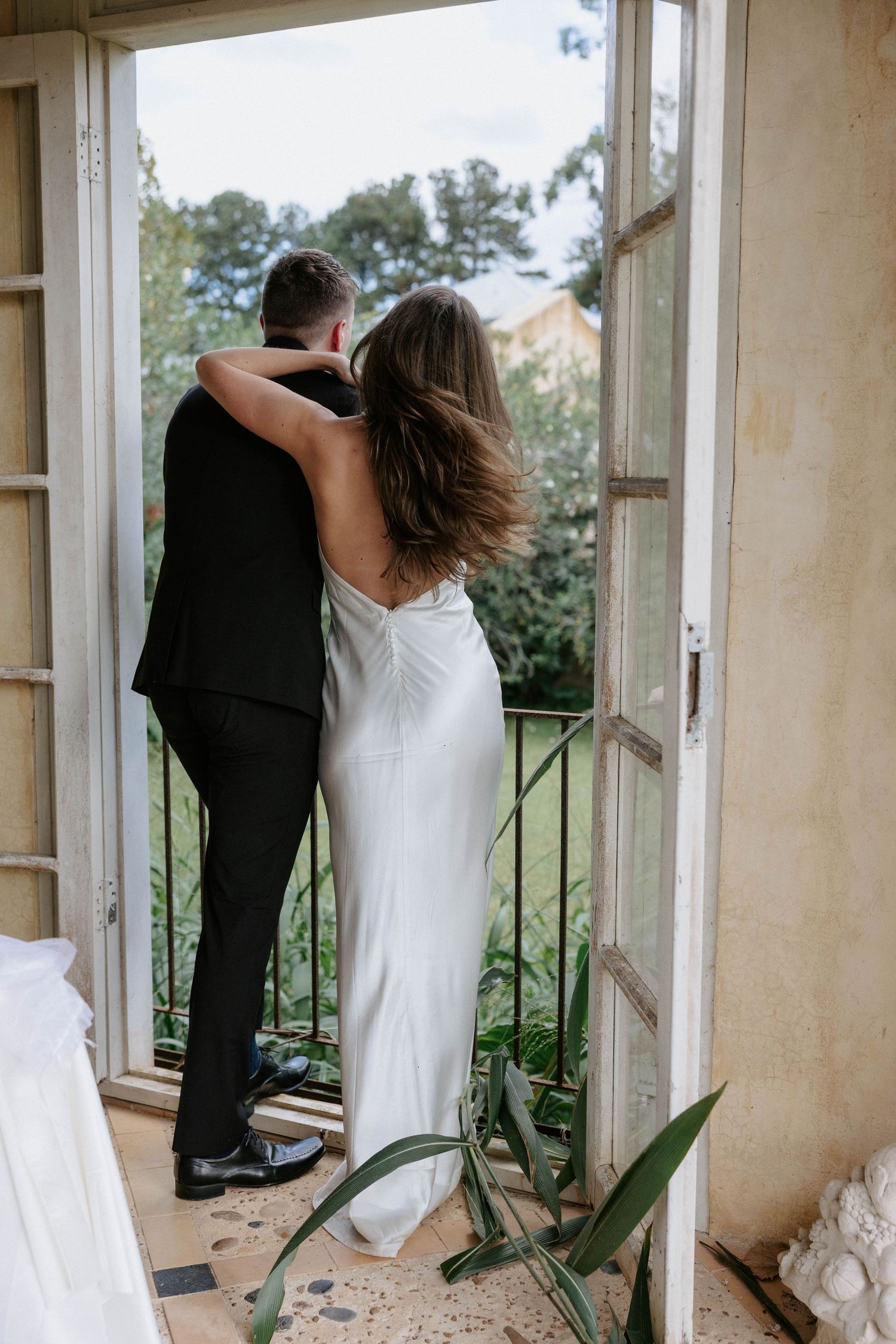 Bride and groom embrace on a balcony doorway, she in a white satin gown, he in a black suit, overlooking lush greenery.