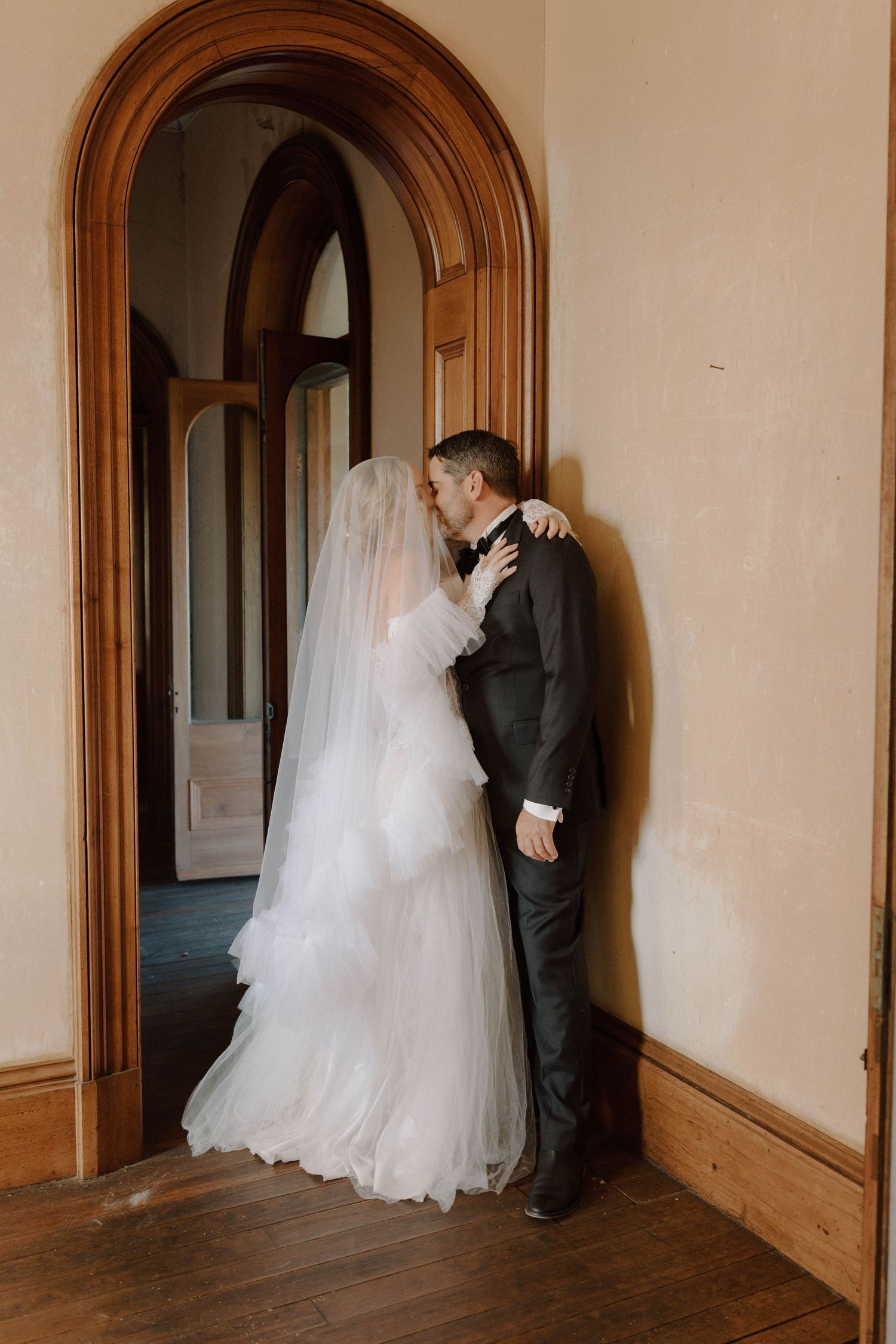 A romantic wedding couple shares an intimate moment in an arched hallway with warm wooden trim.