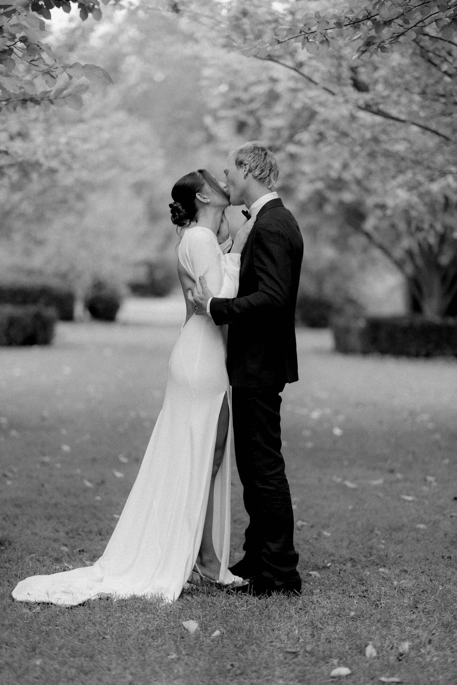 Black and white photo of bride and groom sharing a kiss in a tree-lined garden.