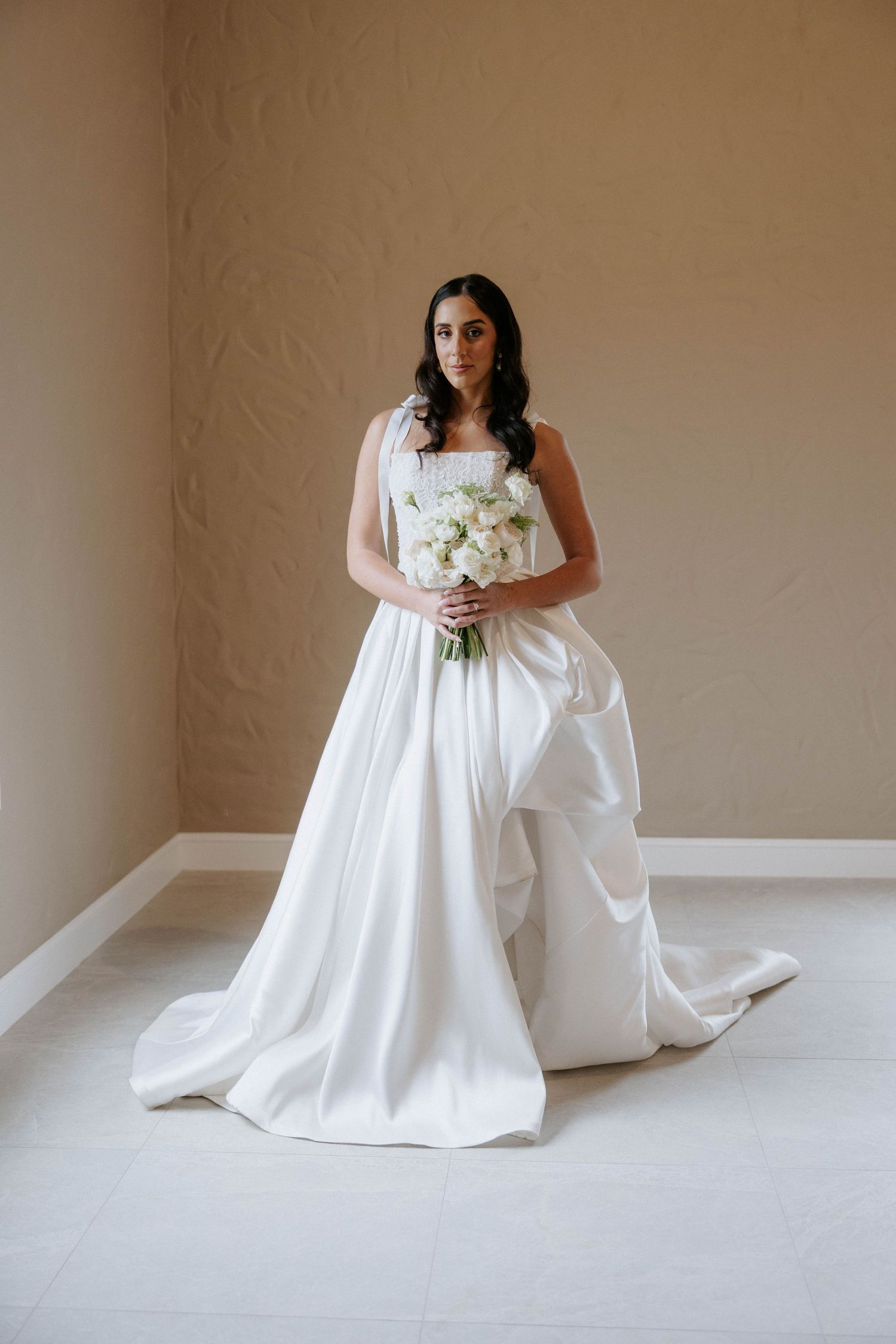 Elegant bride in white satin ball gown with train holds white floral bouquet in neutral toned room.