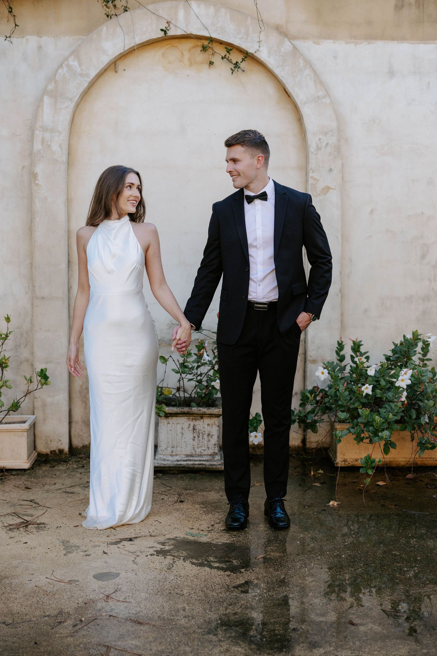 Elegant bride in white halter gown and groom in black tuxedo holding hands outdoors.