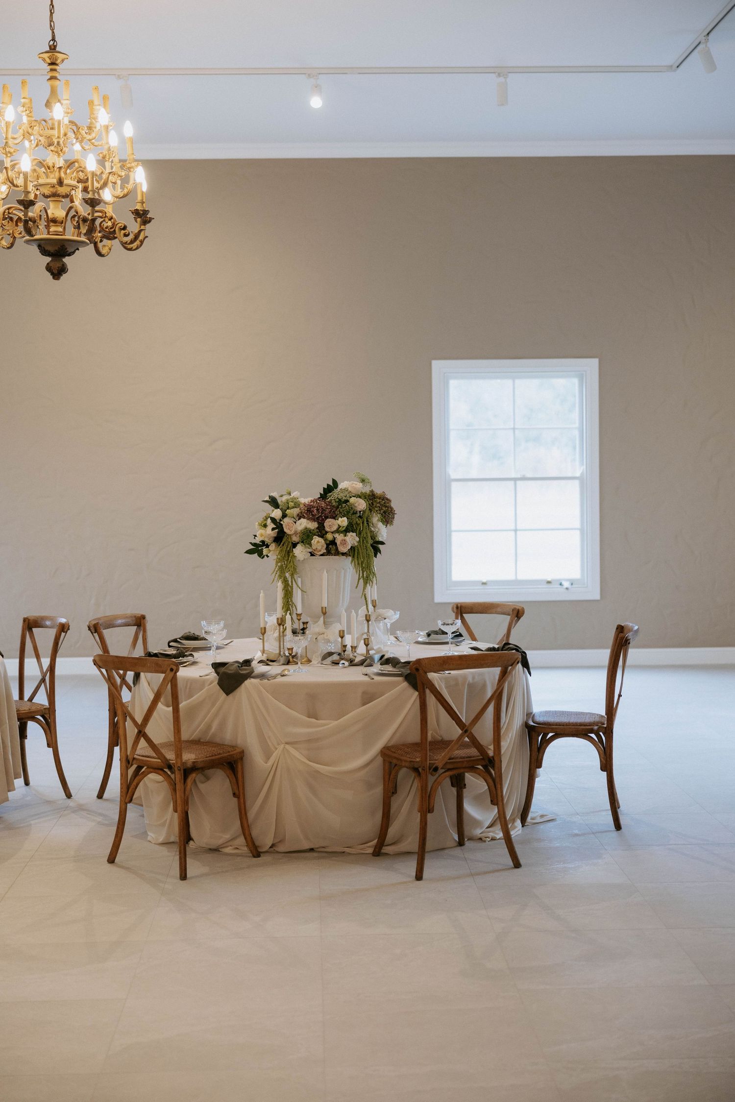 Intimate wedding reception room with wooden cross-back chairs, floral centerpiece, and gold chandelier.