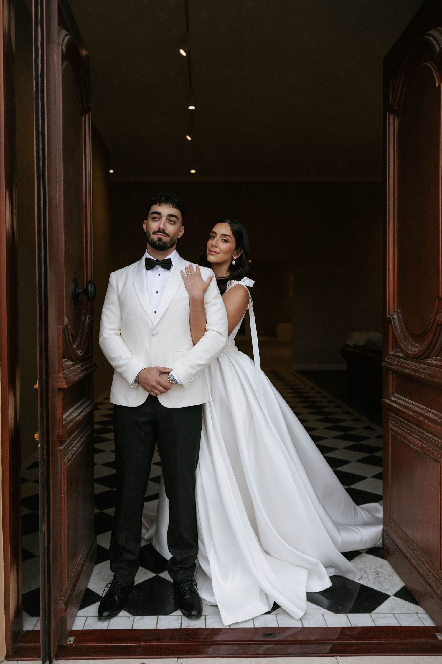 Elegant bride in white gown embraces groom in white tuxedo jacket, posing in doorway with checkered floor.